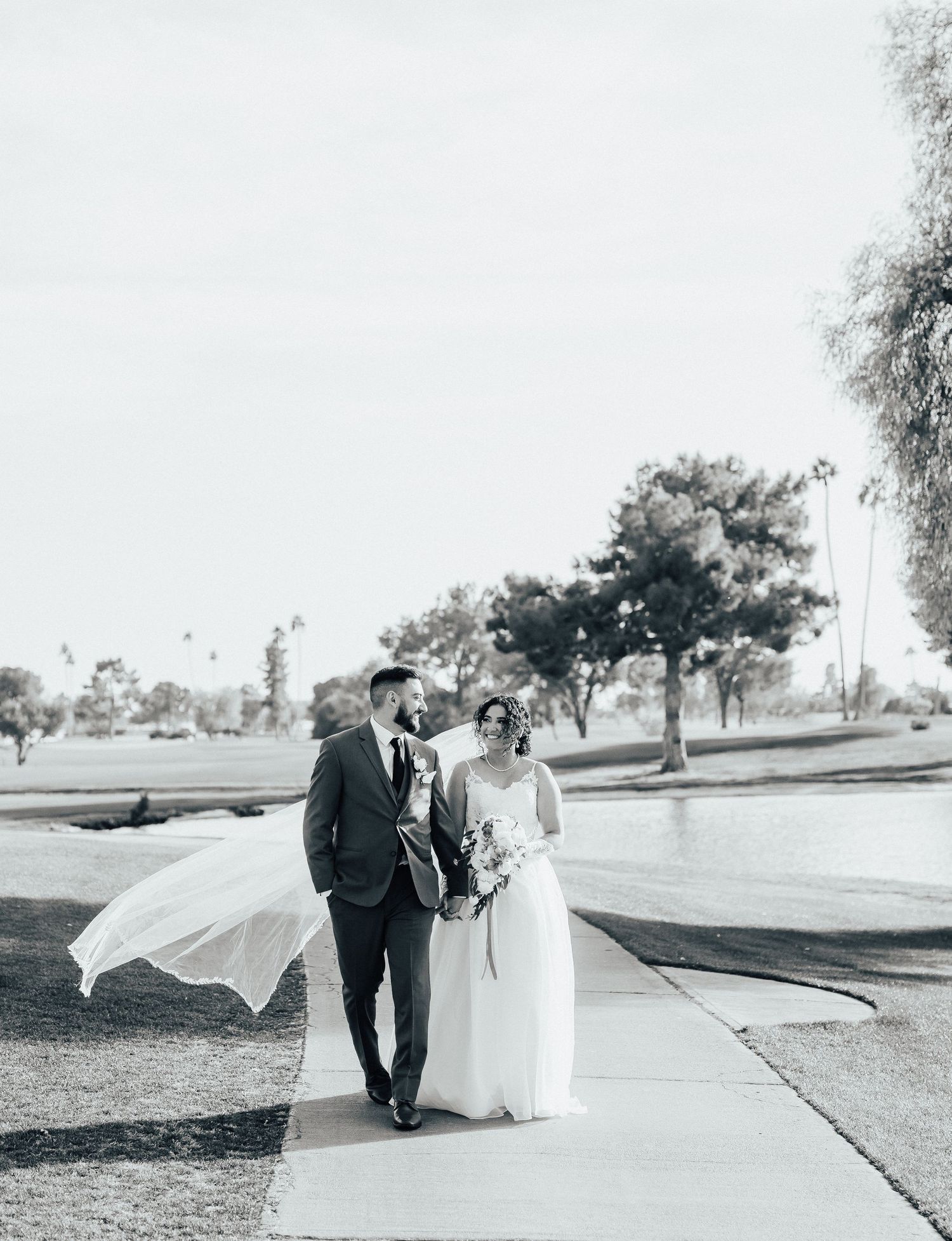 A black and white wedding photo of a couple walking on a path with the bride's veil flowing in the wind.
