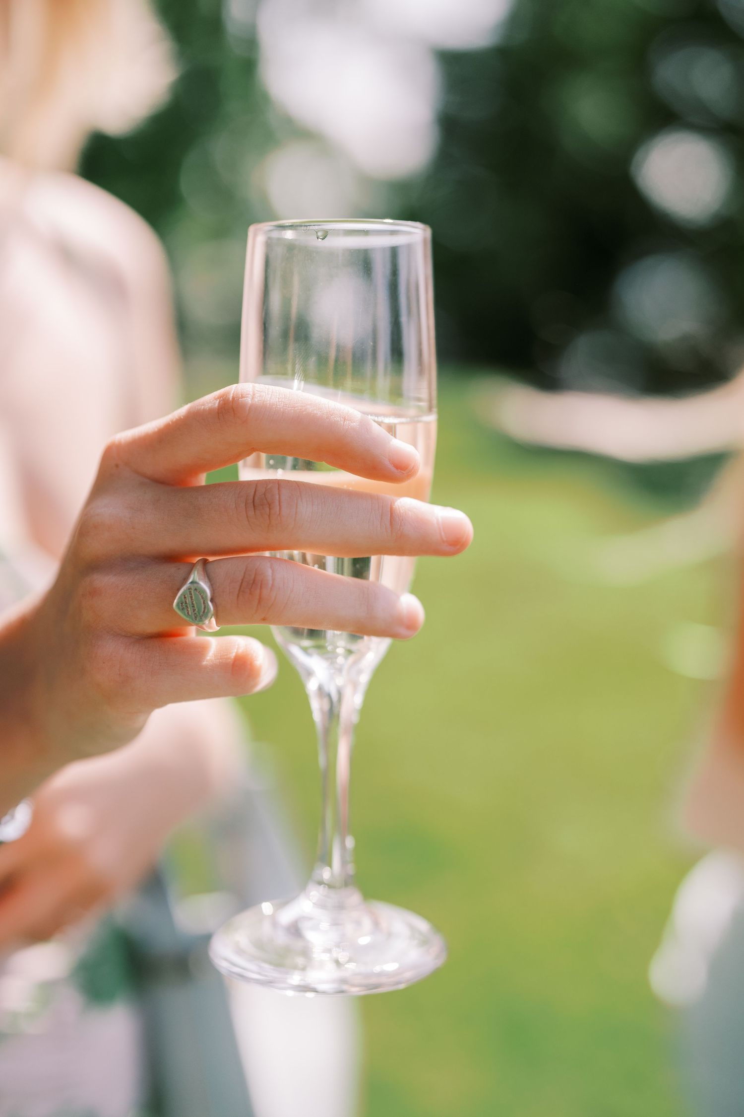 A hand with a turquoise ring holds an elegant champagne flute against a blurred green outdoor background.