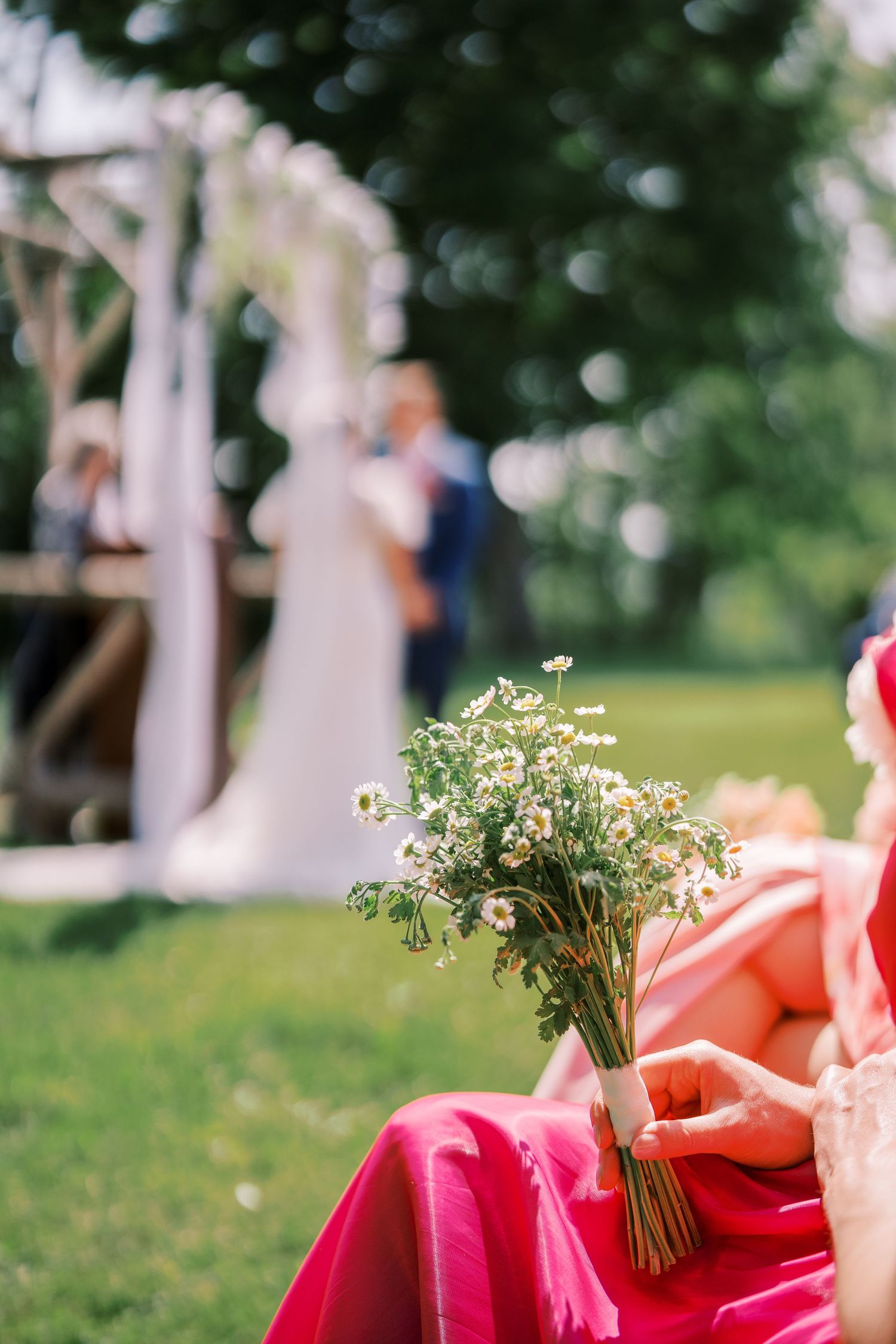 A delicate bouquet of small white wildflowers rests on a red fabric at an outdoor wedding ceremony with a blurred background.