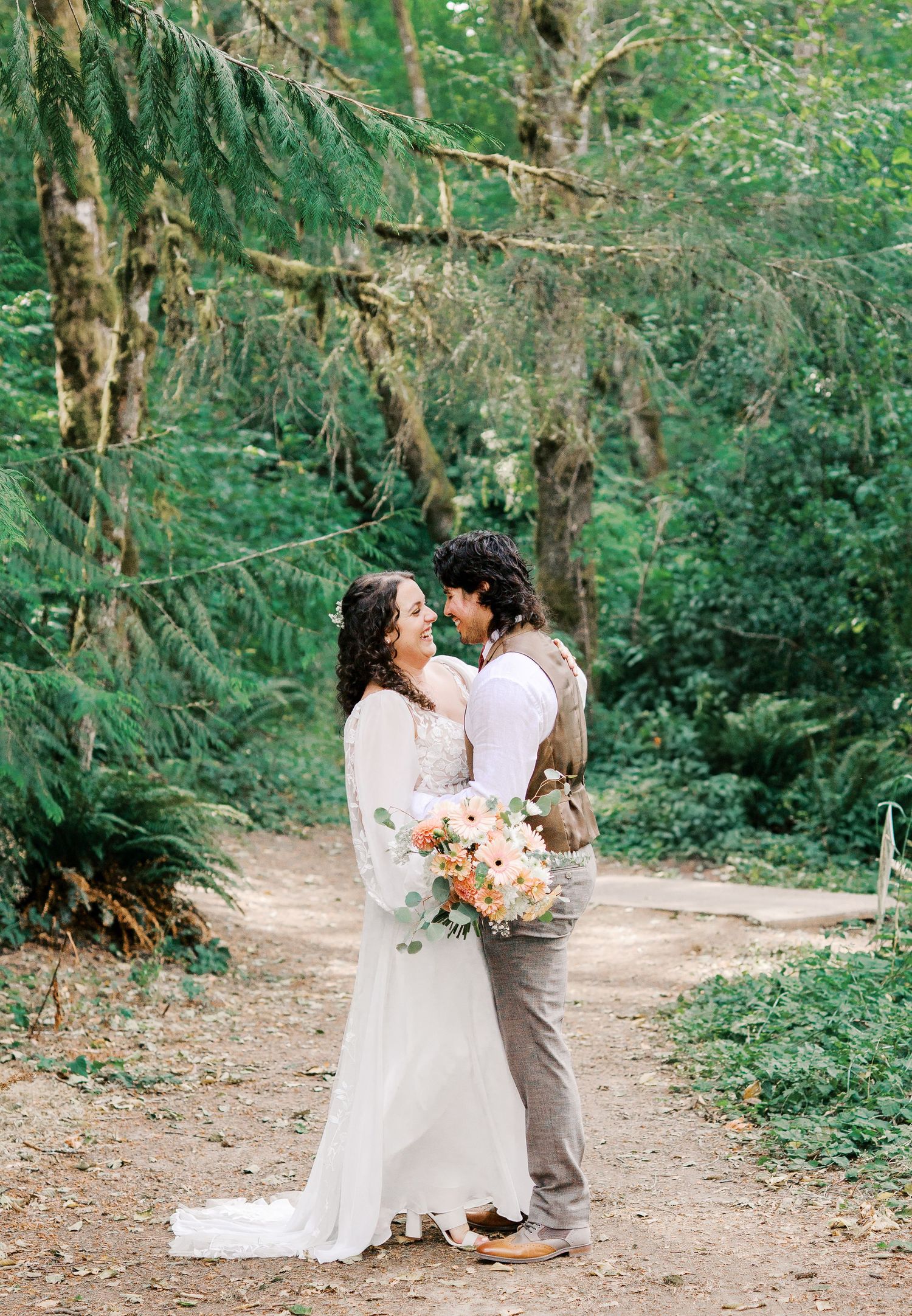 A couple shares an intimate moment on a forest path surrounded by evergreen trees while holding a peach and white bouquet.