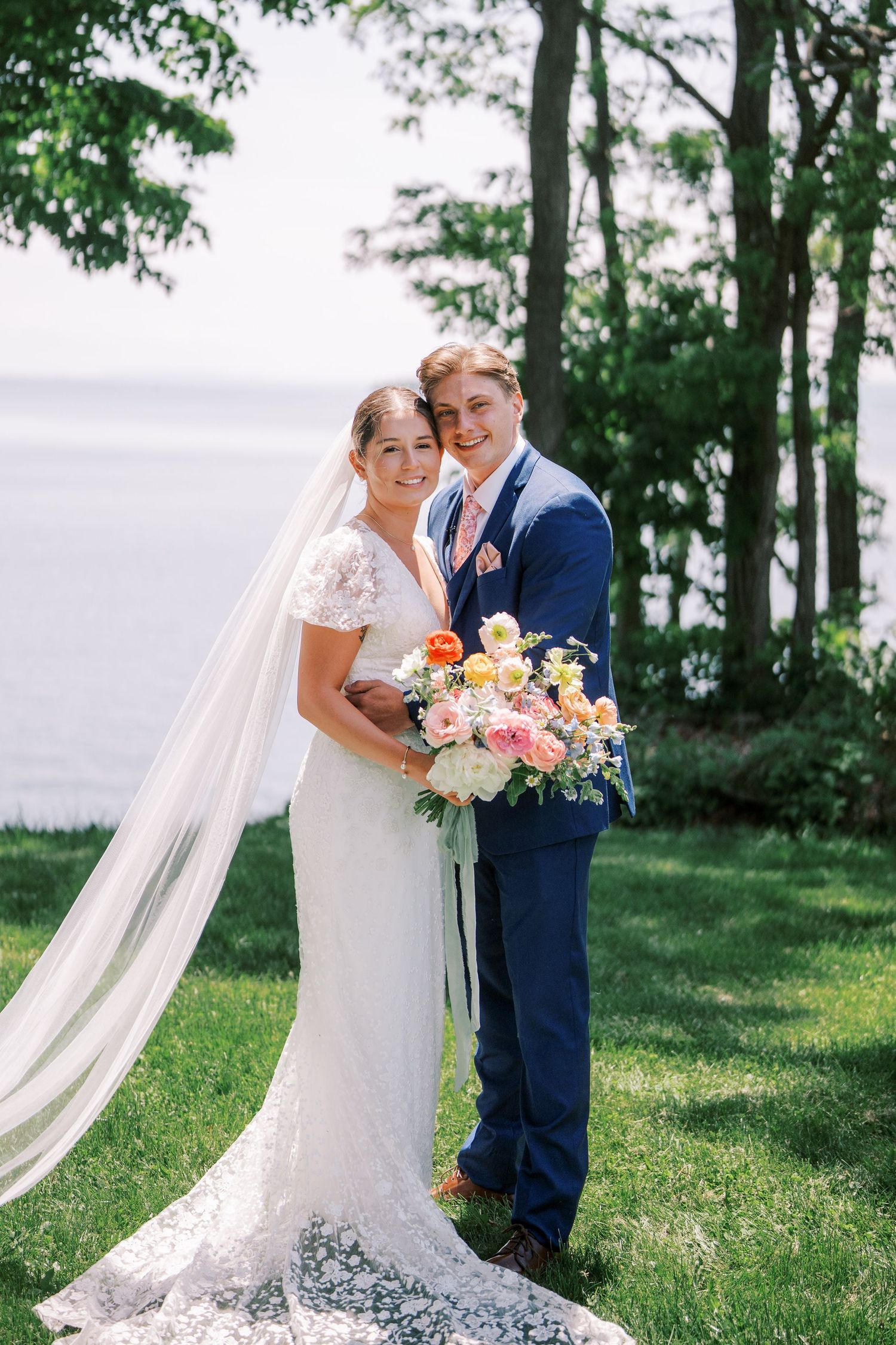Elegant wedding couple poses among trees by the water in a romantic outdoor setting.