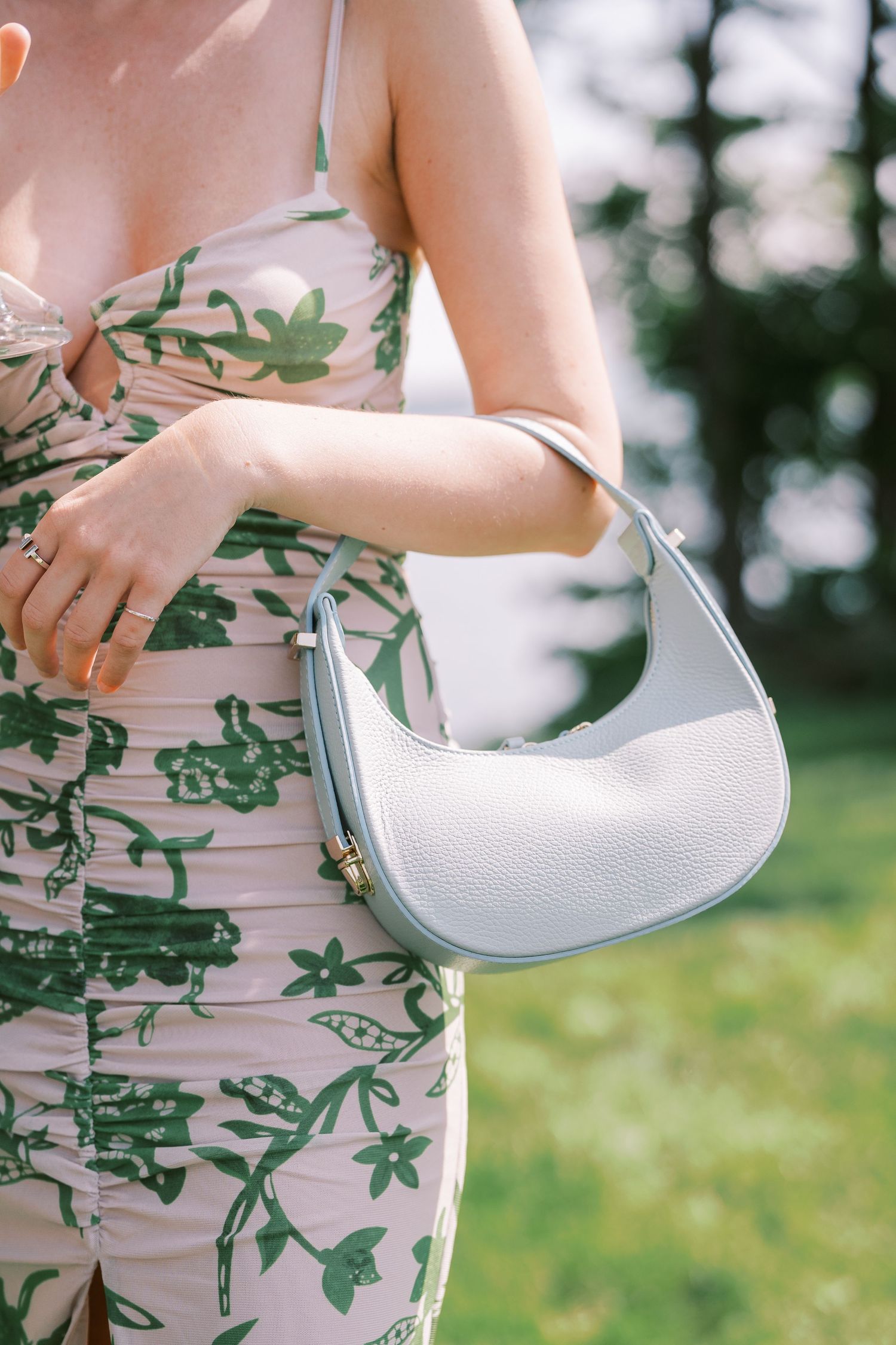 Woman in floral print dress holds a small white crescent-shaped handbag against blurred outdoor background.