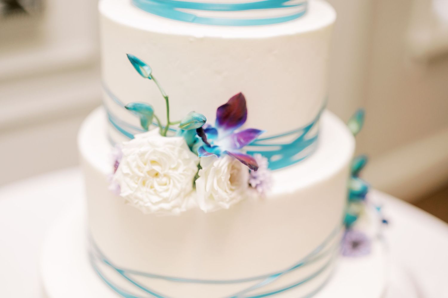 A white tiered wedding cake decorated with turquoise ribbons and white and purple flowers.