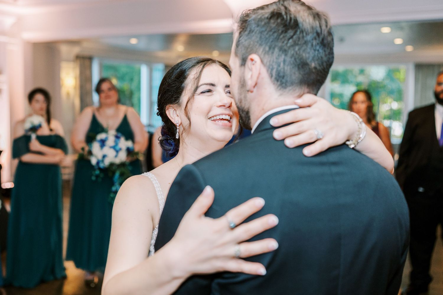 A couple embraces and laughs during their first dance at a wedding reception while guests watch in the background.