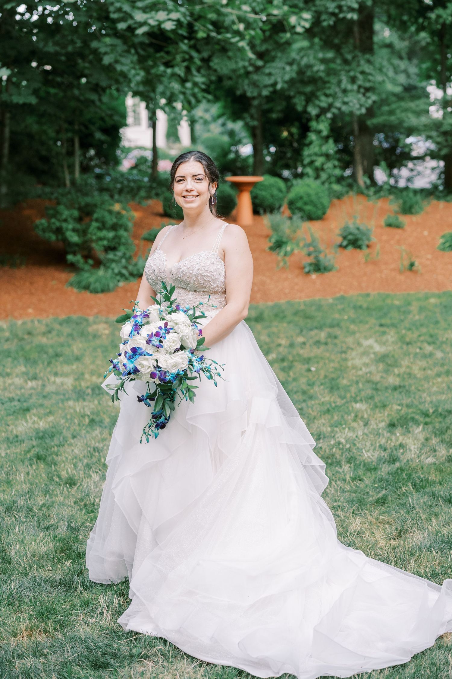 A bride in a flowing white gown with layered tulle skirt holds a blue and white floral bouquet in an outdoor garden setting.