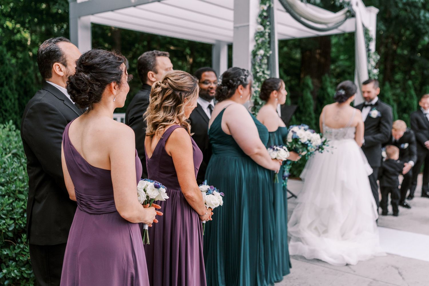 Bridesmaids in purple and teal dresses stand together at an outdoor wedding ceremony.