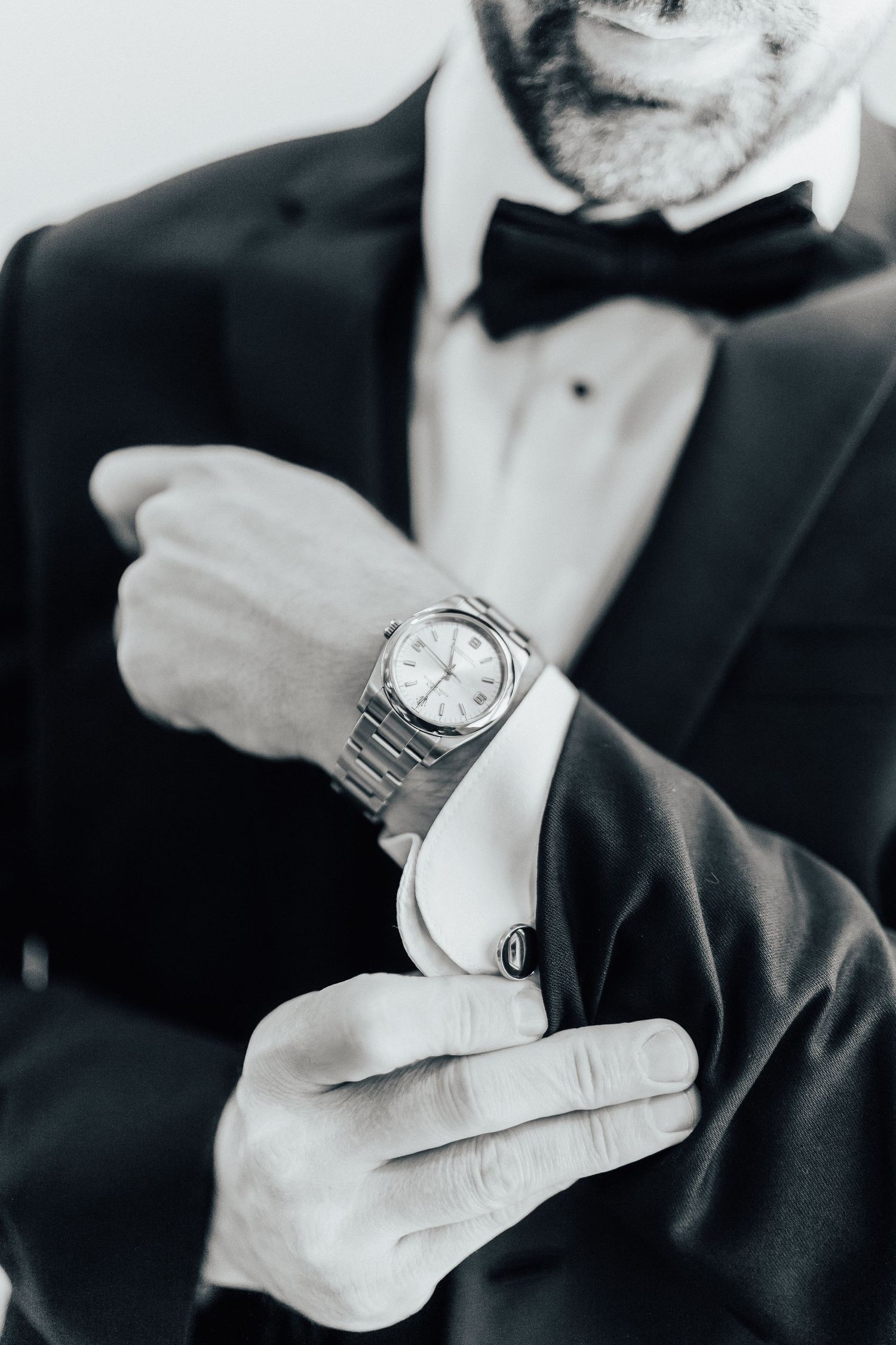 Black and white photo showing hands adjusting a luxury watch with a tuxedo and bowtie in the background.