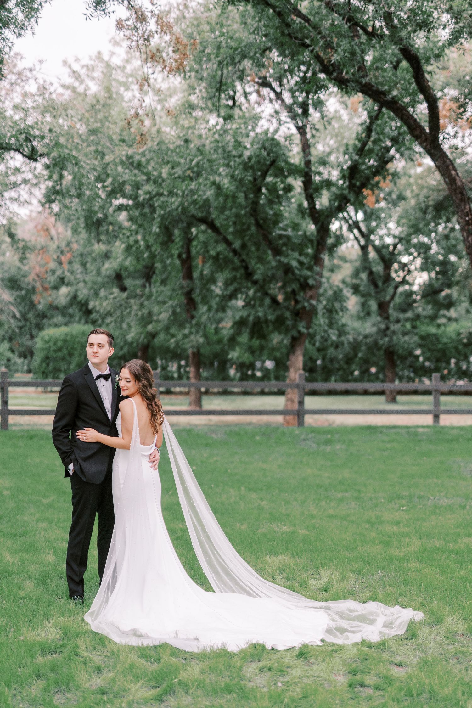 A wedding couple walks through a serene park setting with a long flowing white veil trailing behind them.