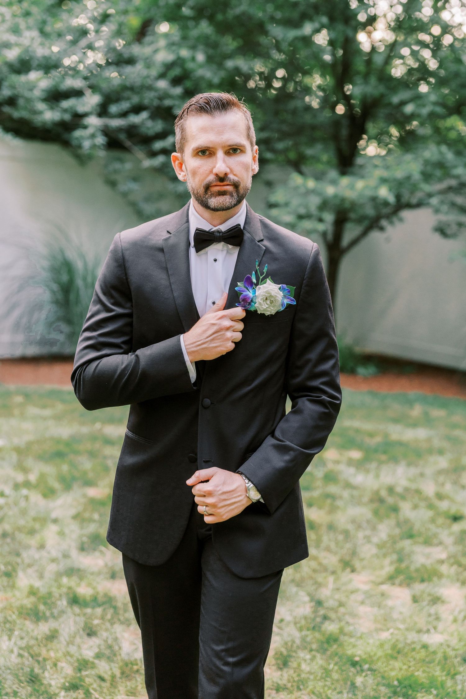 Wedding attire featuring classic black tuxedo with bow tie and delicate blue floral boutonniere in outdoor garden setting.