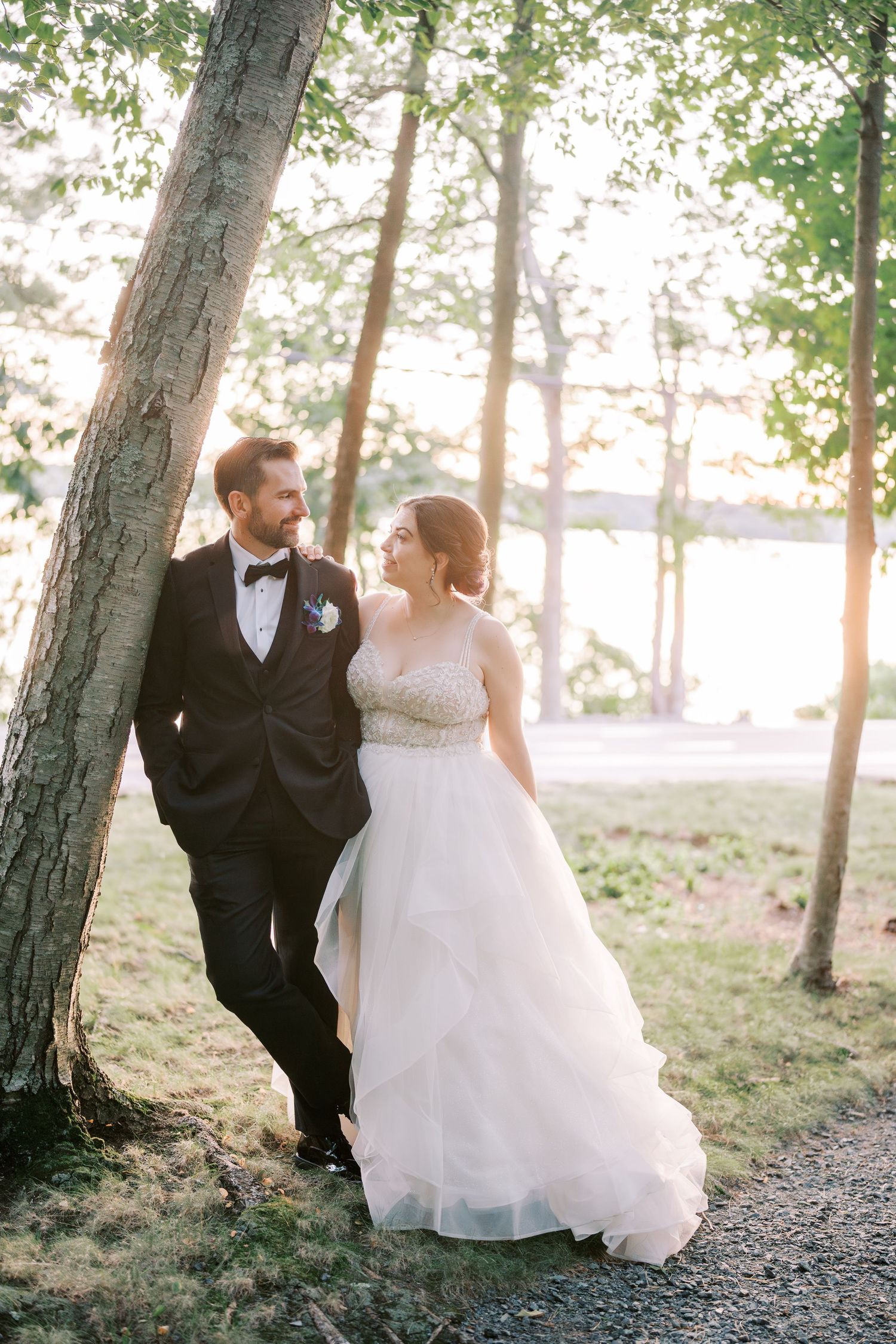 Newlywed couple shares intimate moment under trees at sunset with lake in background.