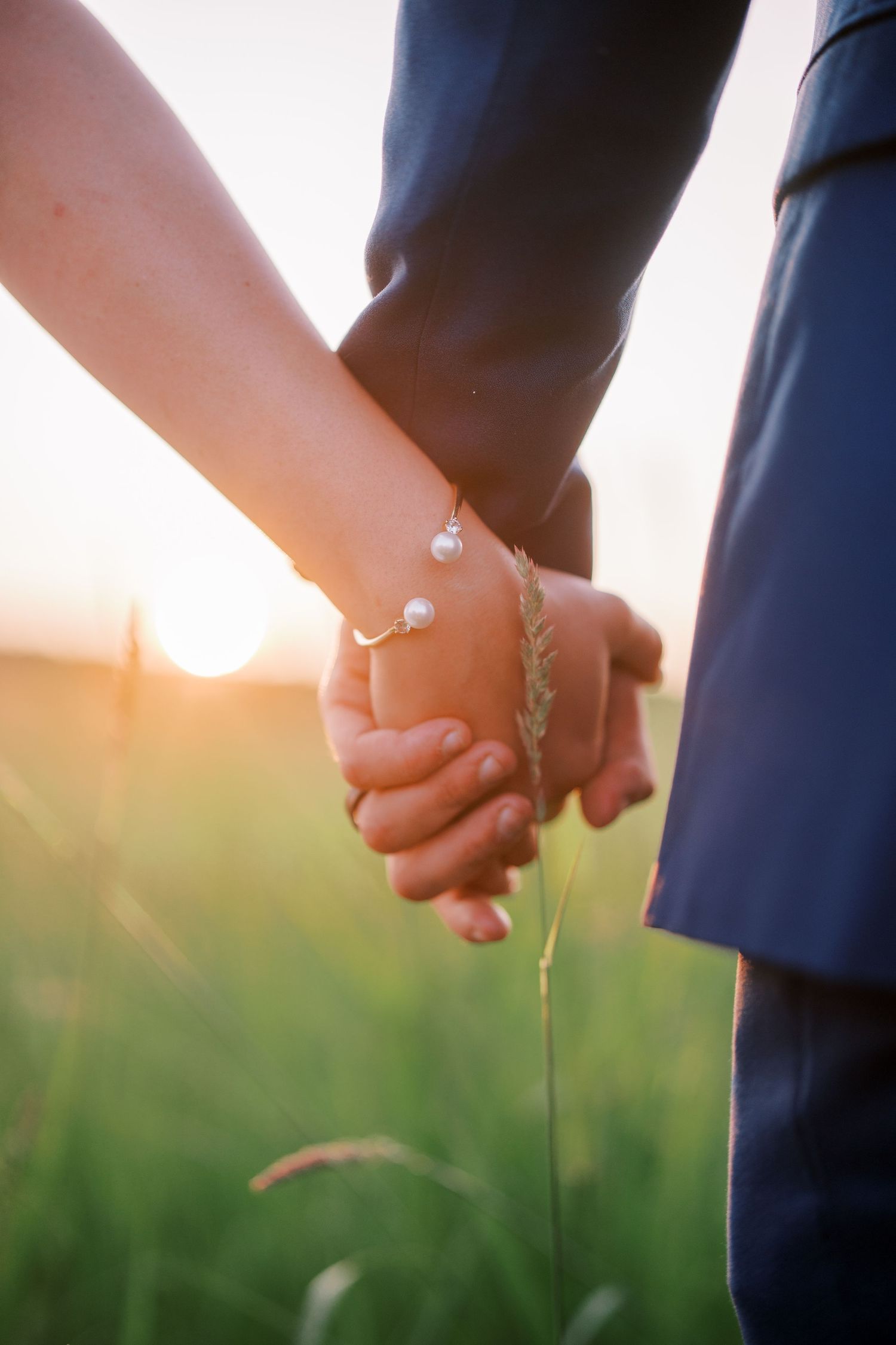 Two people holding hands in a sunlit field with grass, one wearing a delicate pearl bracelet.