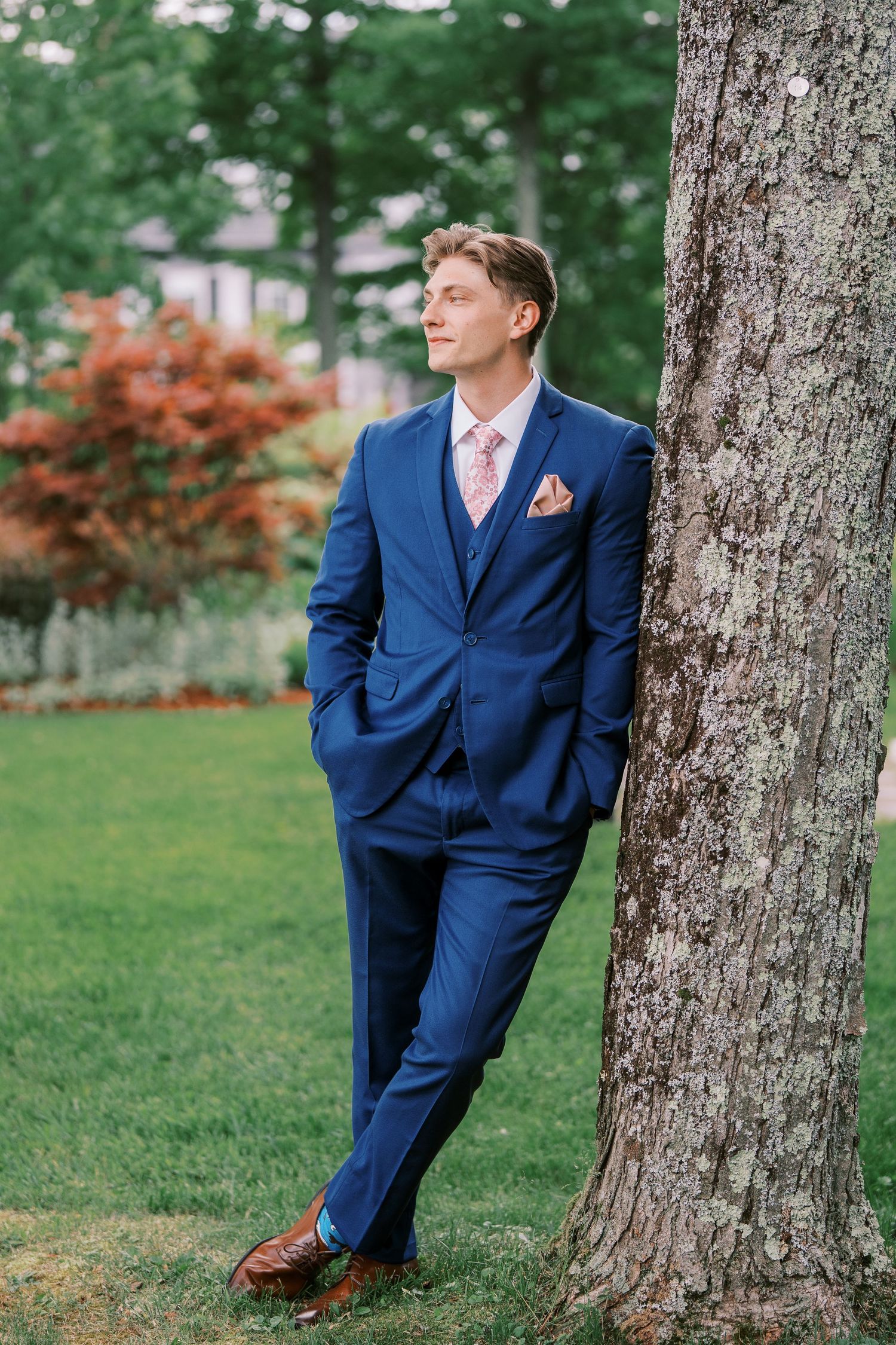 A person wearing a navy blue suit poses elegantly against a tree trunk in an outdoor garden setting with fall foliage.