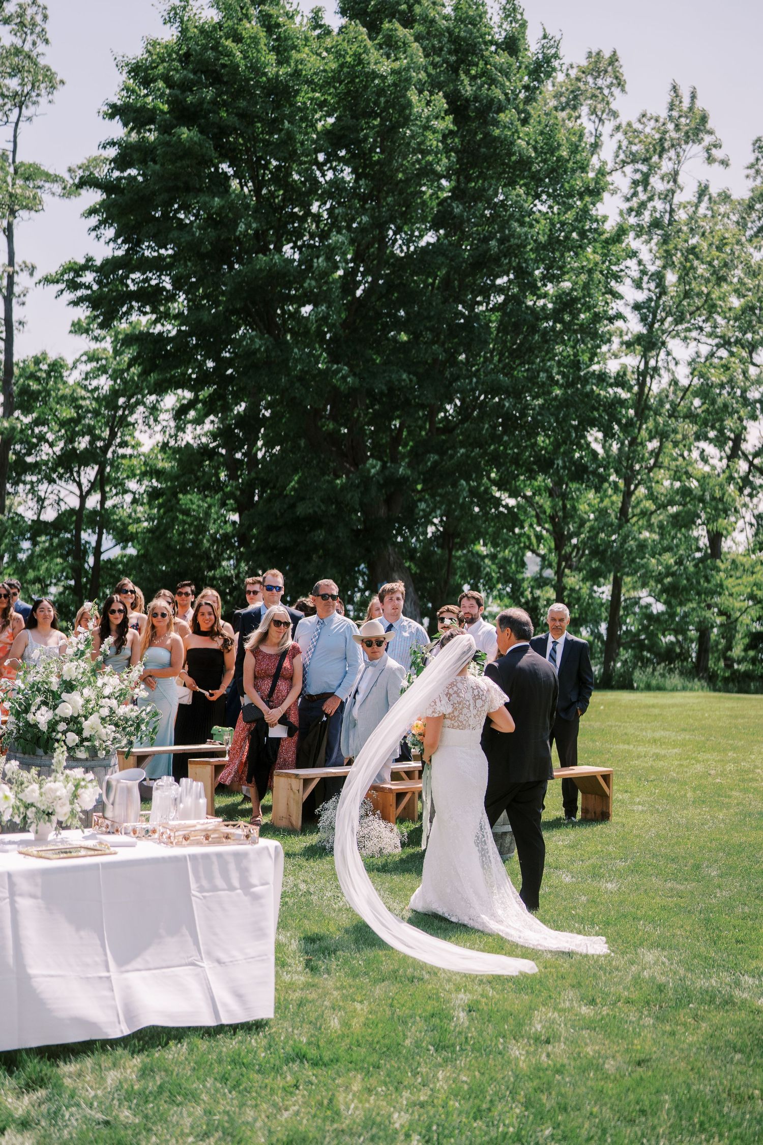 A festive wedding ceremony takes place outdoors on a grassy hilltop with guests gathering to celebrate in a scenic natural setting.