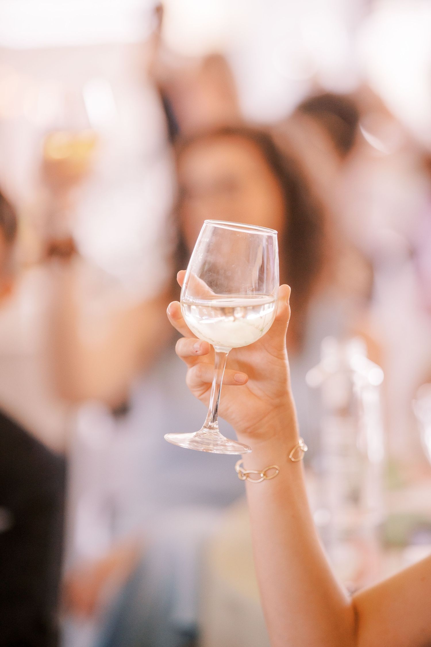 A hand raises a delicate wine glass filled with white wine in a celebratory toast at an event.