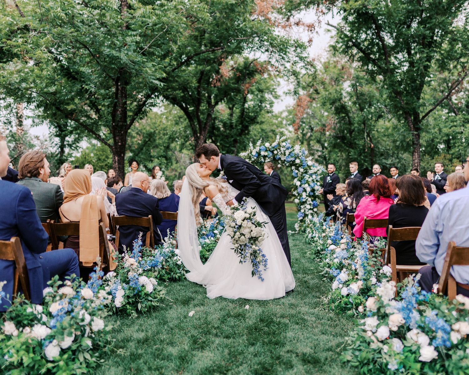 Couple shares romantic moment during outdoor garden wedding ceremony surrounded by blue floral arrangements and seated guests.