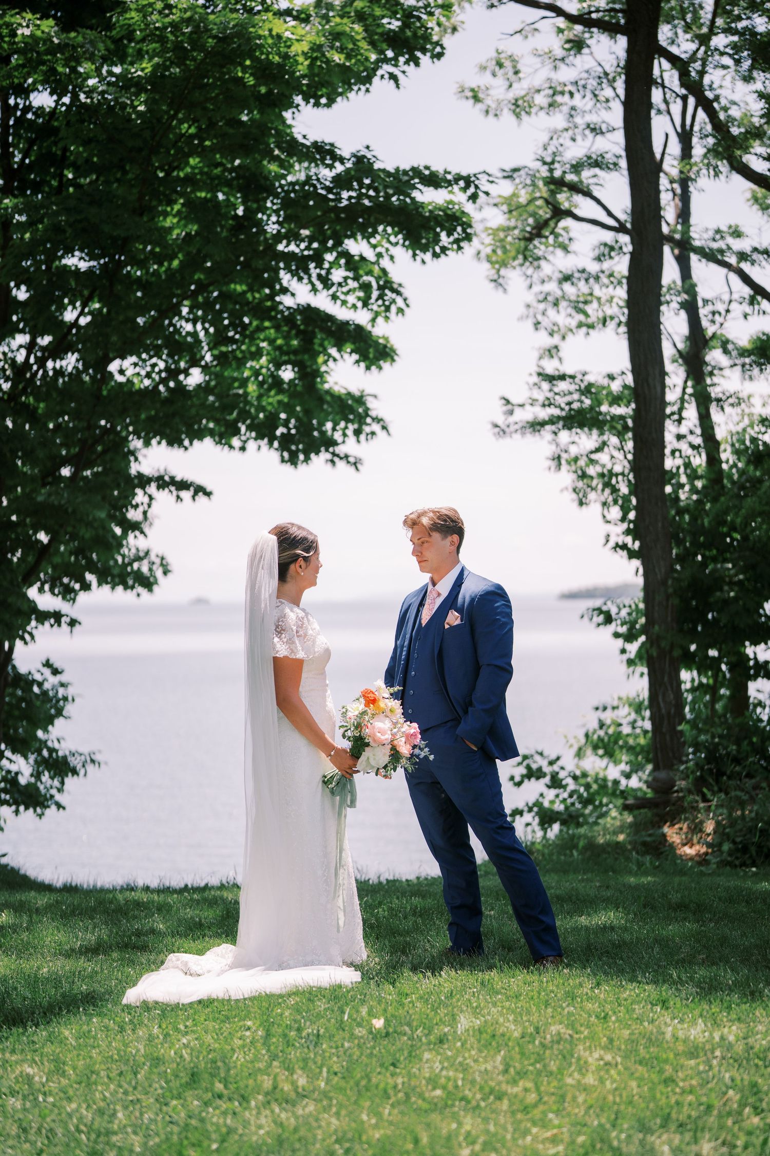 A couple stands together on a grassy hill overlooking a lake, surrounded by trees on a sunny summer day.