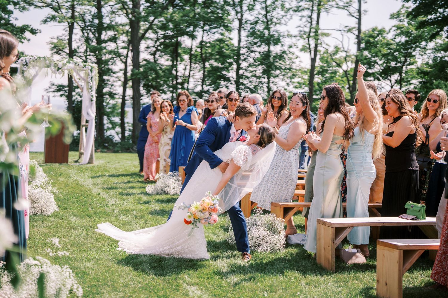 Joyful outdoor wedding ceremony with guests celebrating on a grassy hill surrounded by tall green trees.