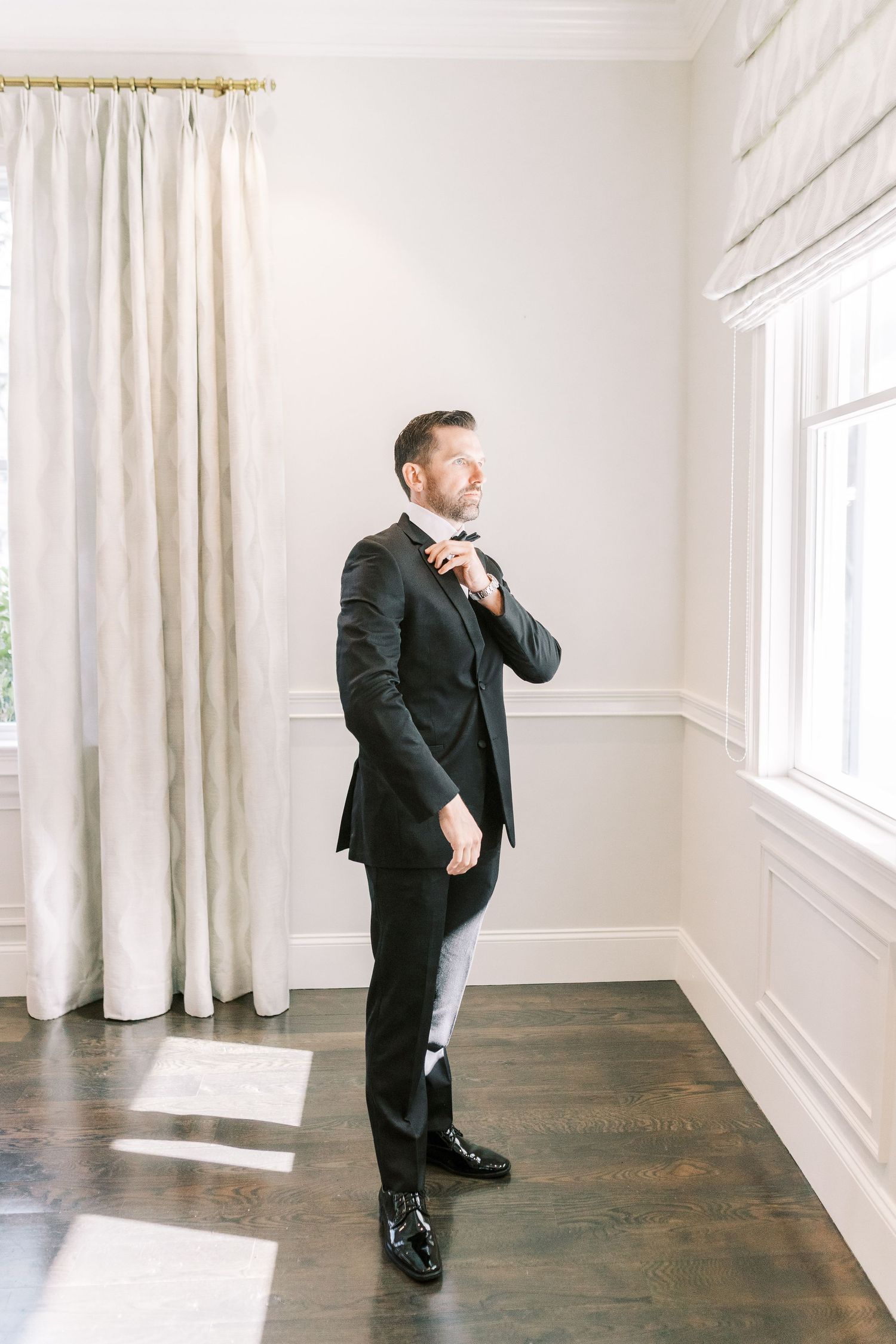 A person in a formal black suit and tie adjusts their collar while standing in a bright room with white curtains and walls.