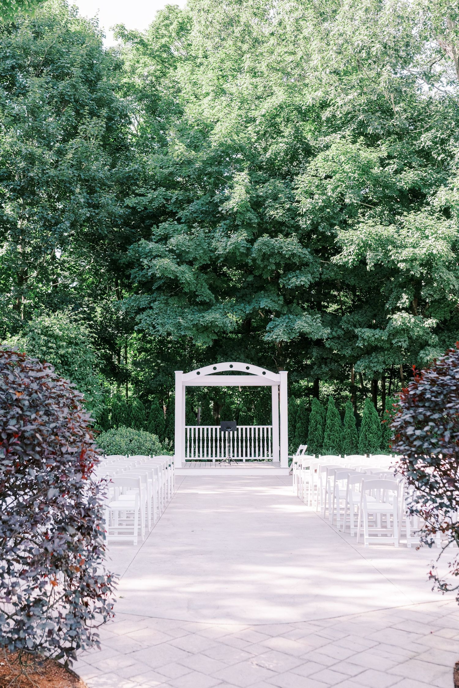 White garden wedding arbor with rows of chairs set up on stone patio surrounded by lush green trees and burgundy foliage.