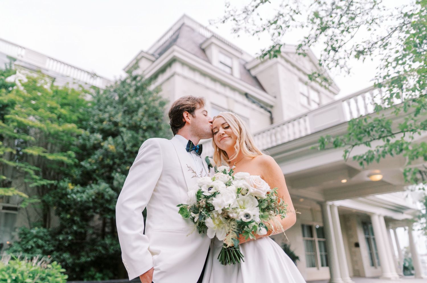 A romantic wedding couple embraces outdoors in front of an elegant historic mansion with a white floral bouquet.