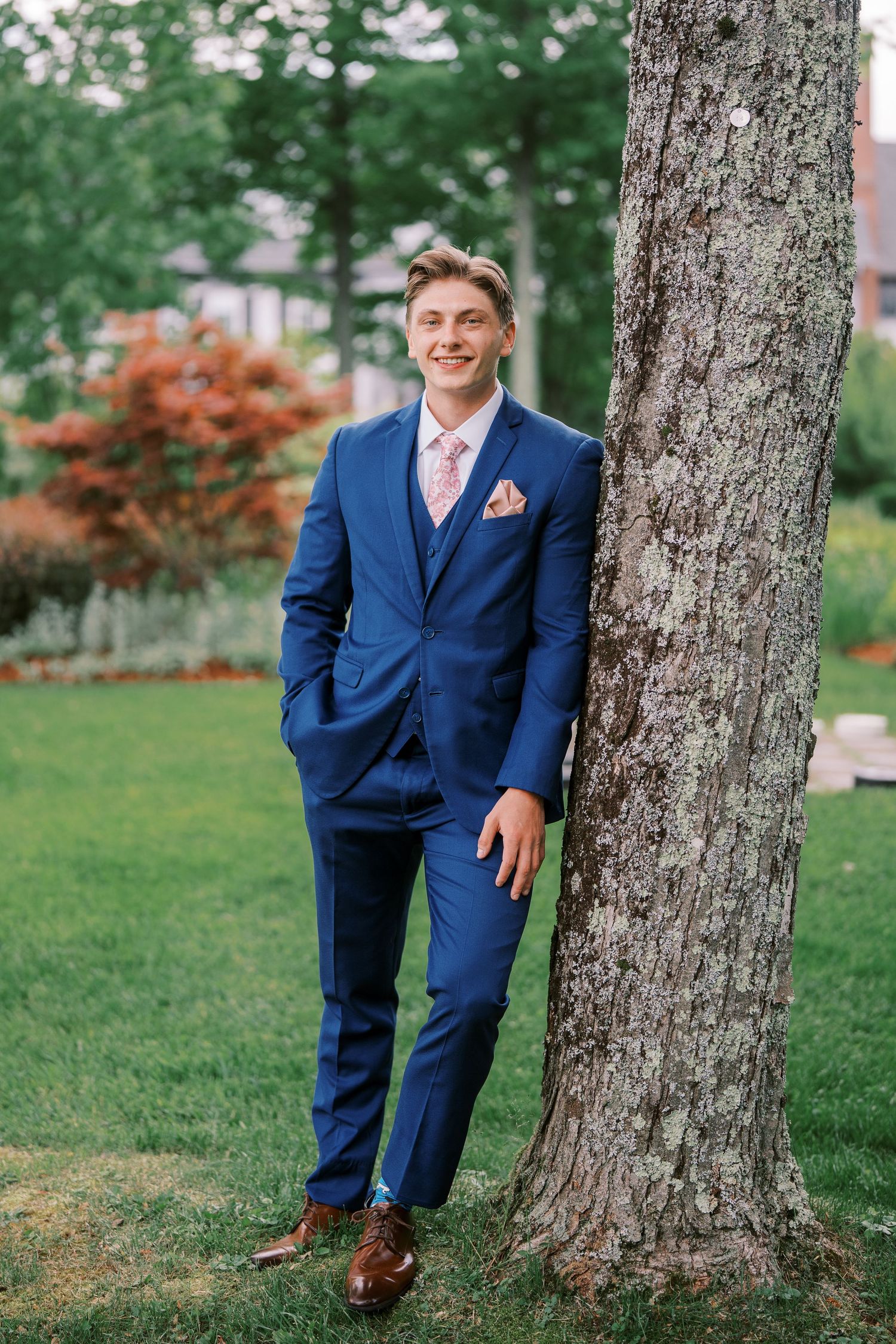 A person in a navy blue suit with pink tie and pocket square poses casually against a large tree trunk in a garden setting.
