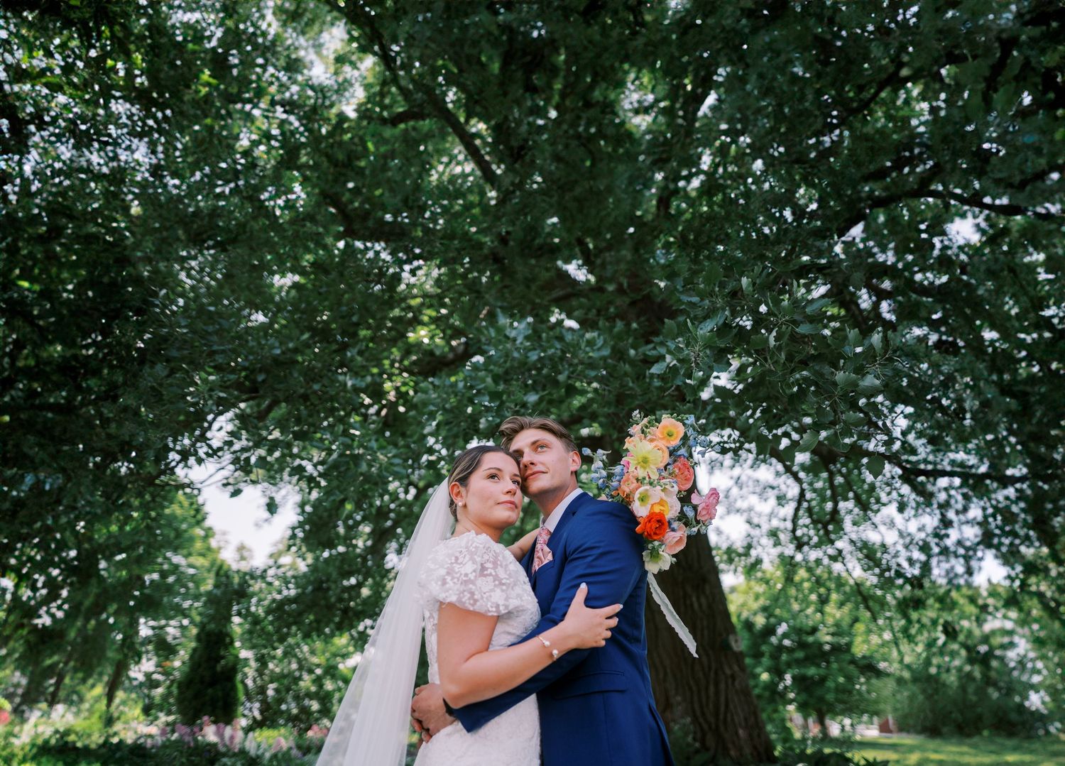 A newlywed couple embraces under a lush green canopy of trees during an outdoor wedding celebration.