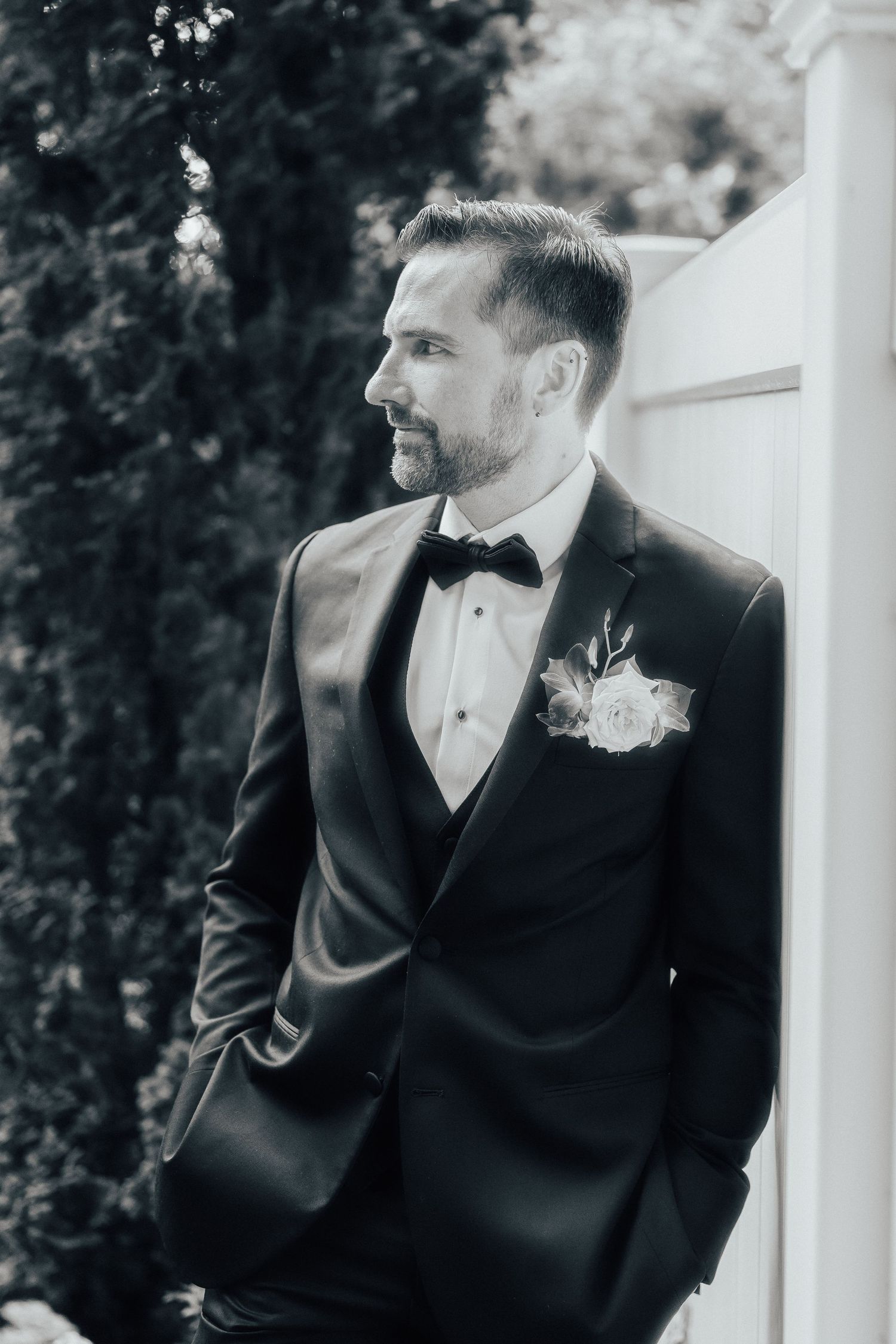 Classic black and white wedding portraits showing groom in tuxedo with boutonniere standing against light backdrop.