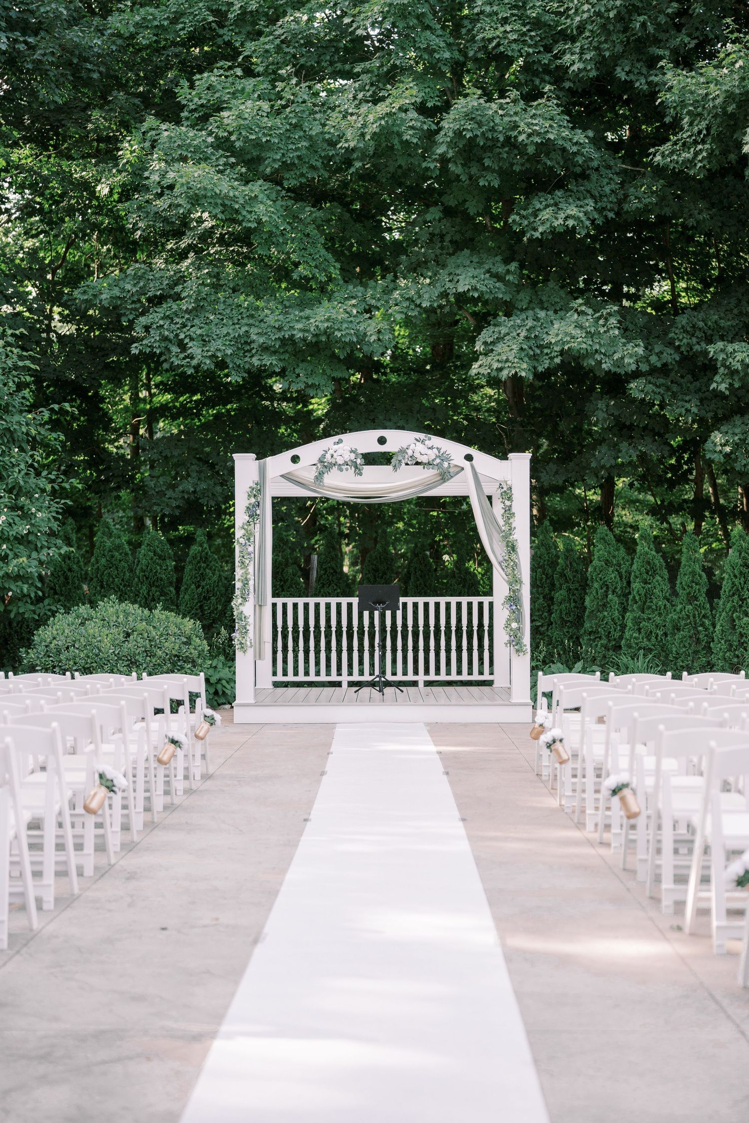 An elegant outdoor wedding ceremony setup with white chairs and a decorative arch in a wooded garden setting.