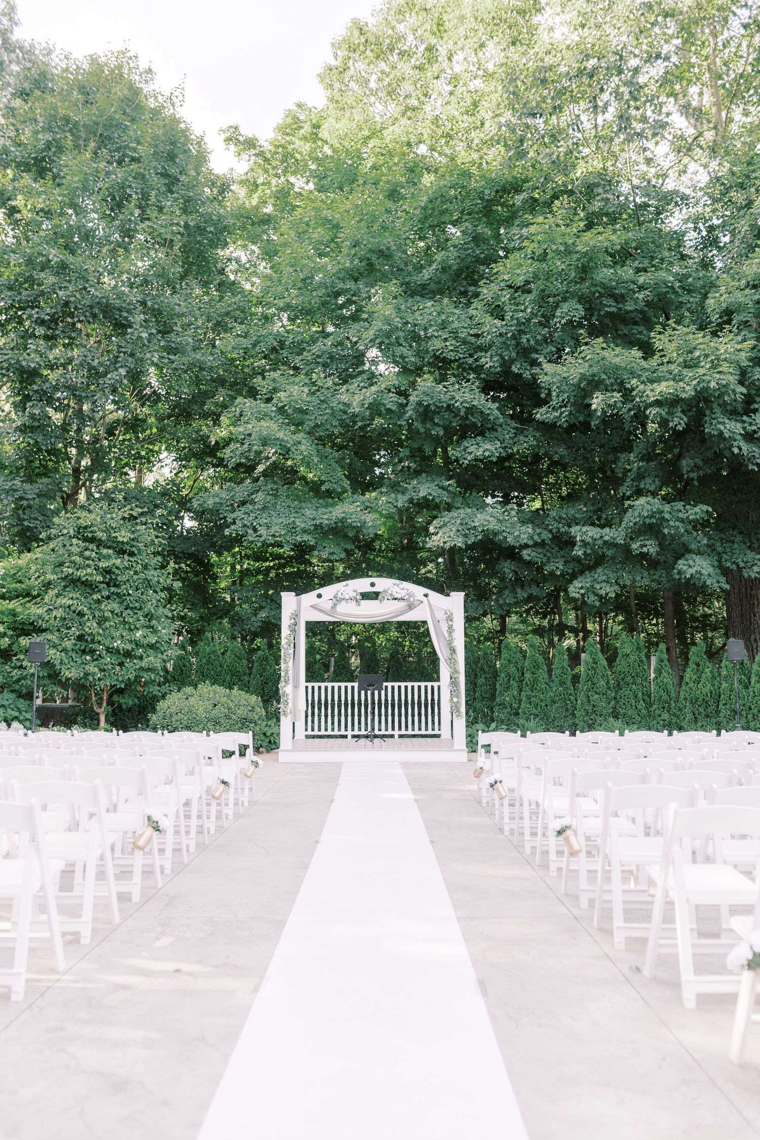 A panoramic view of an outdoor wedding ceremony setup with white chairs and arch surrounded by lush green trees.