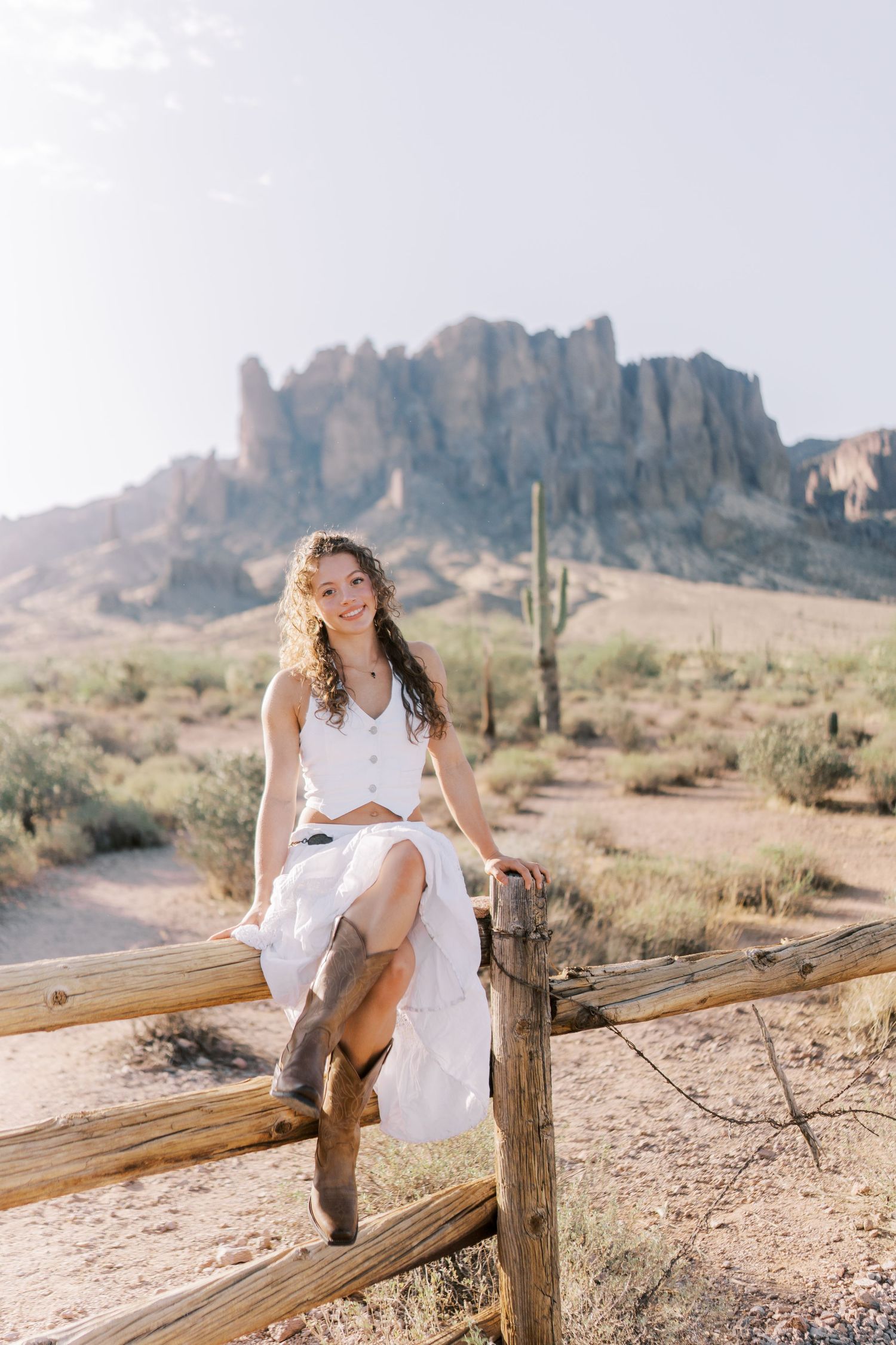 Woman in white dress sitting on wooden fence with Superstition Mountains and saguaro cactus in Arizona desert background.