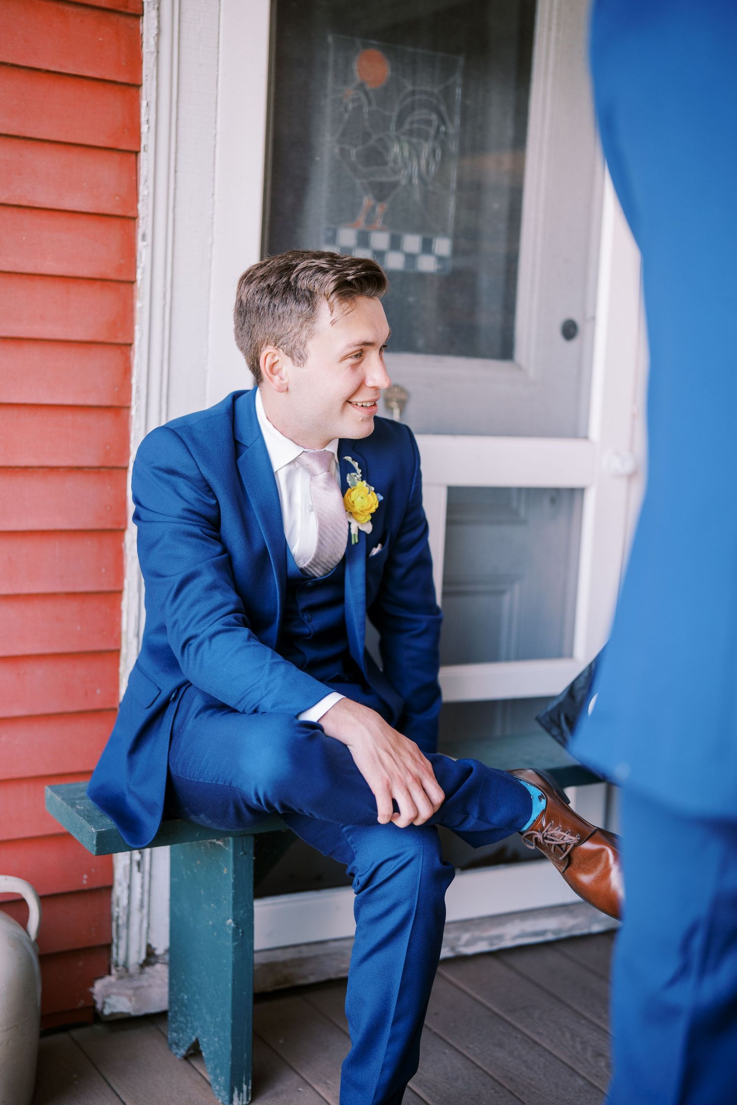 A person in a navy blue suit sits on a covered porch beside a coral-colored house exterior.
