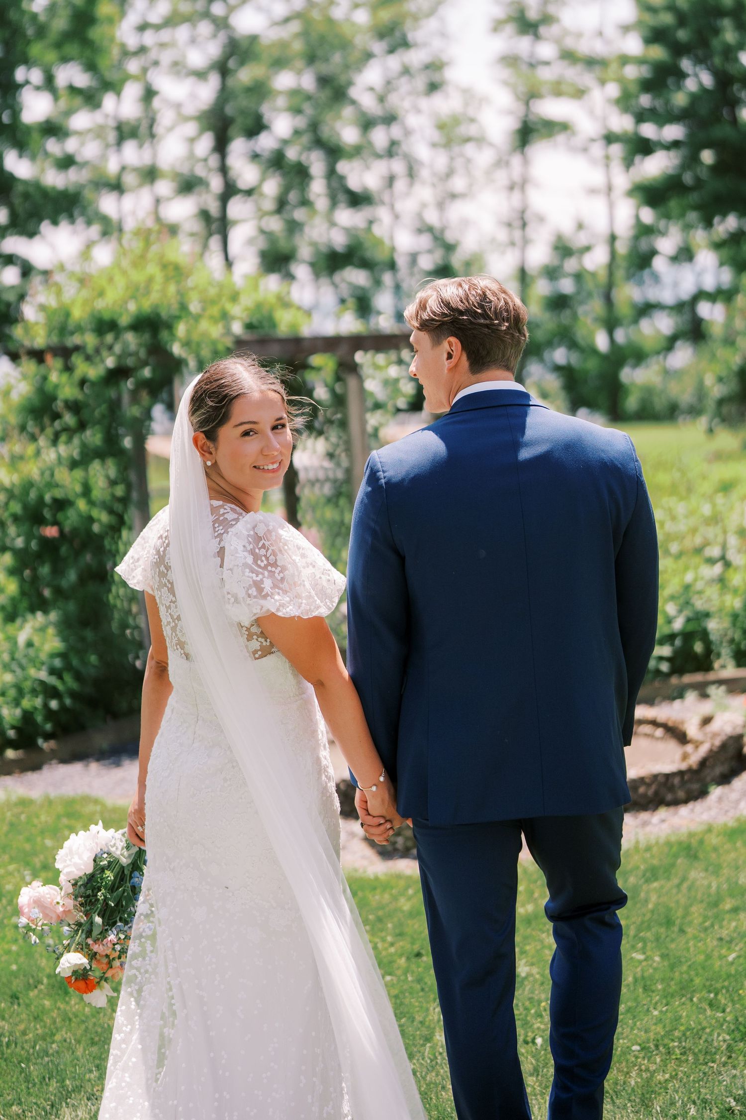 A bride in a white lace gown looks back while holding hands with her groom in a navy suit during an outdoor garden ceremony.