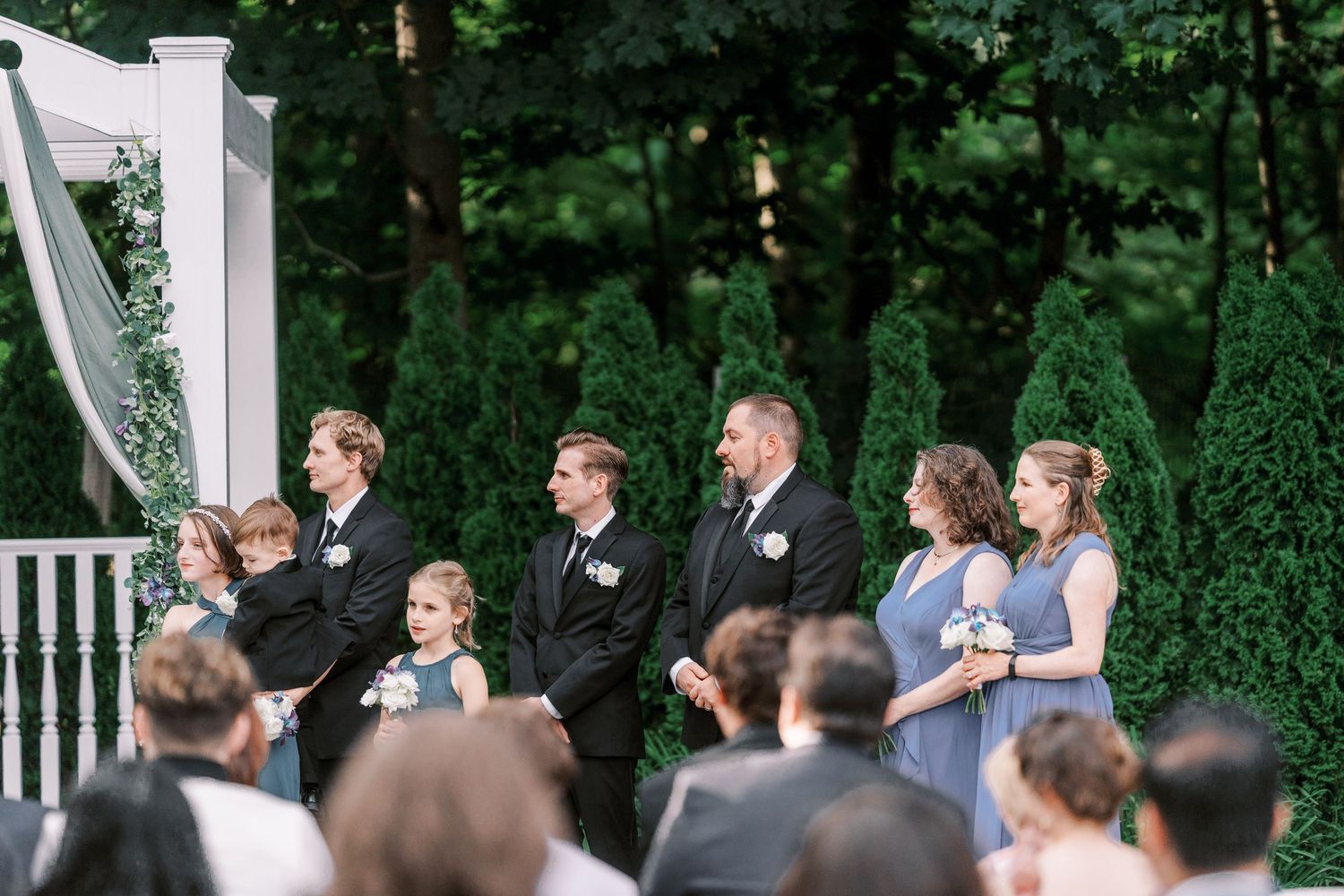 A wedding party stands at an outdoor ceremony gazebo surrounded by lush green trees and guests.