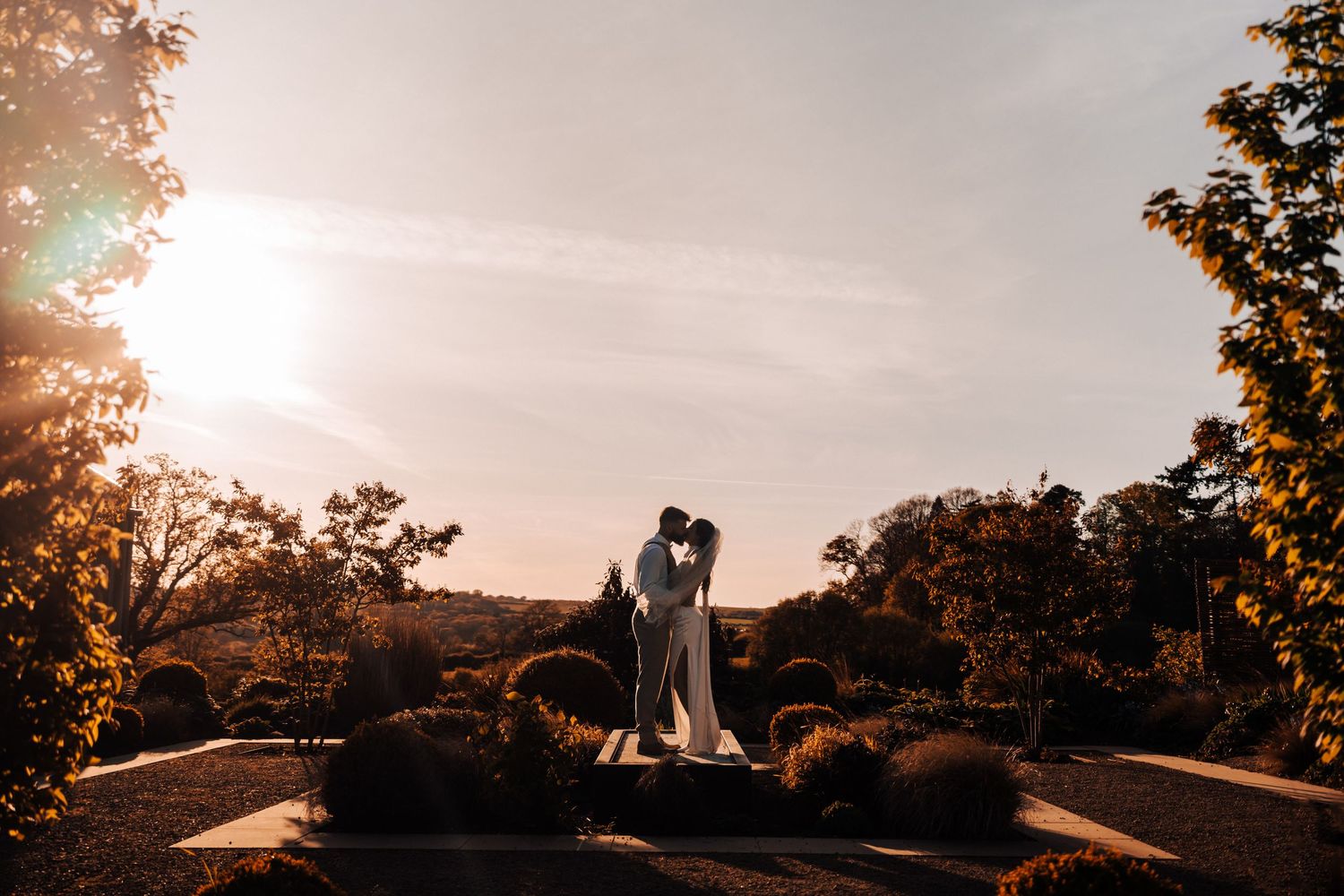 Two silhouettes stand close together on a hilltop at sunset with trees and gardens in the background.
