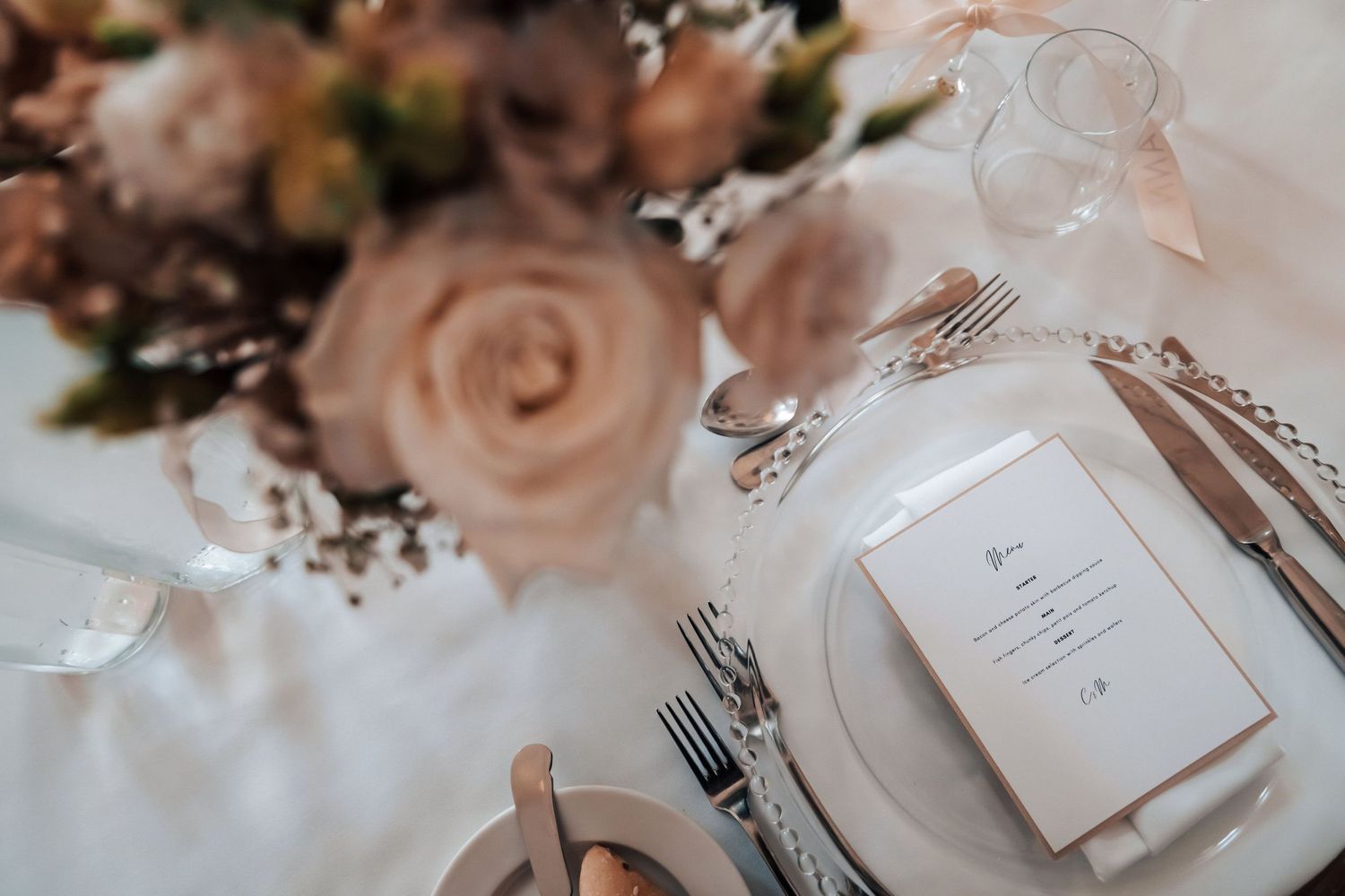 Place settings with elegant dinnerware, pink roses, and menu cards create a romantic wedding reception tablescape.