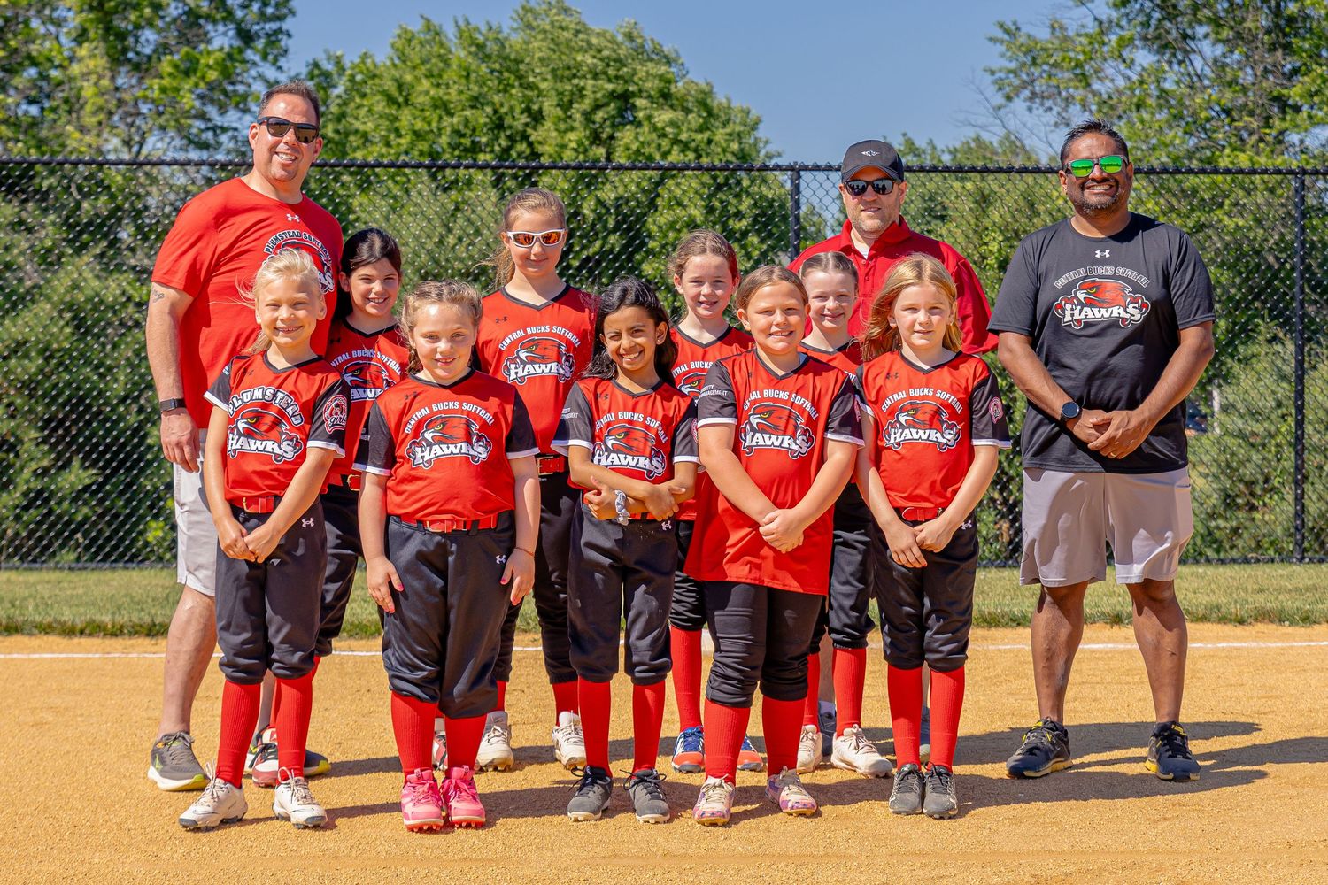 Young girls softball team in red uniforms pose with coaches for a team photo on a sunny day at the baseball field.