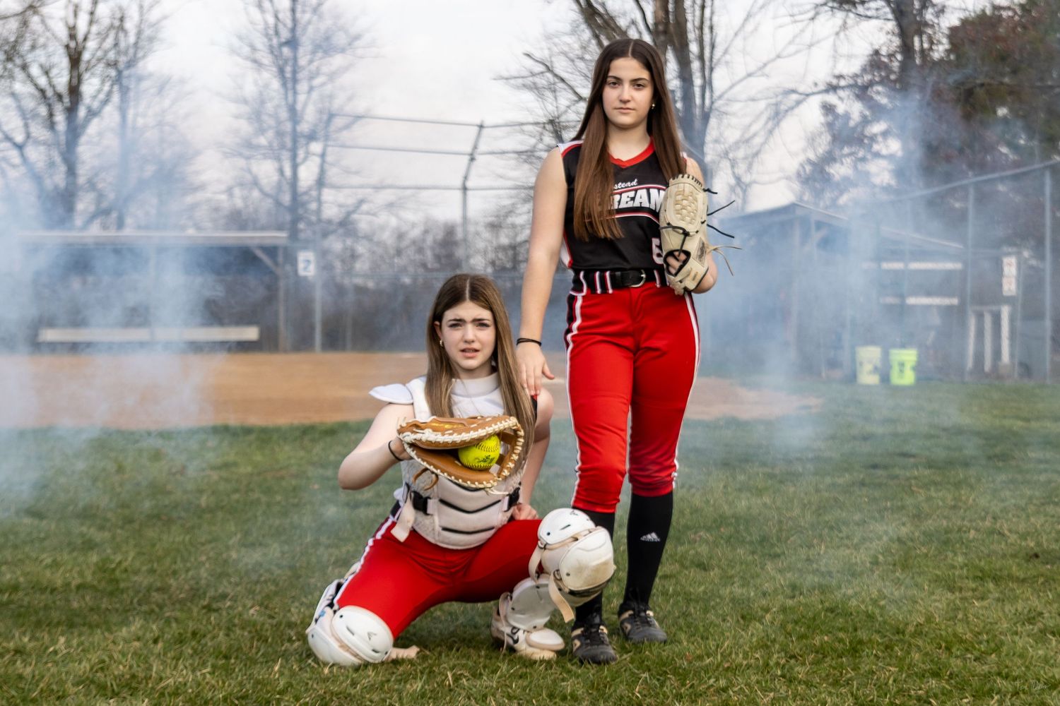Two female softball players pose on a field, one kneeling with a glove, wearing red and white uniforms.
