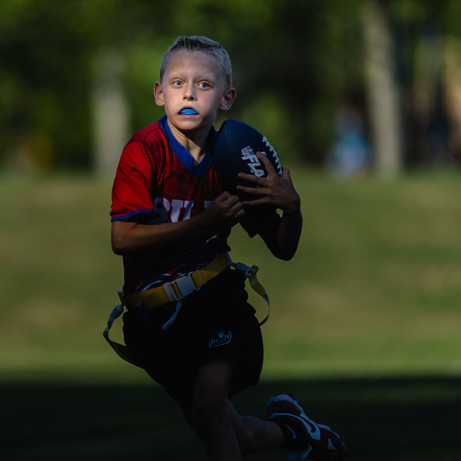 Young boy in red jersey runs with football during flag football game, wearing blue mouthguard.