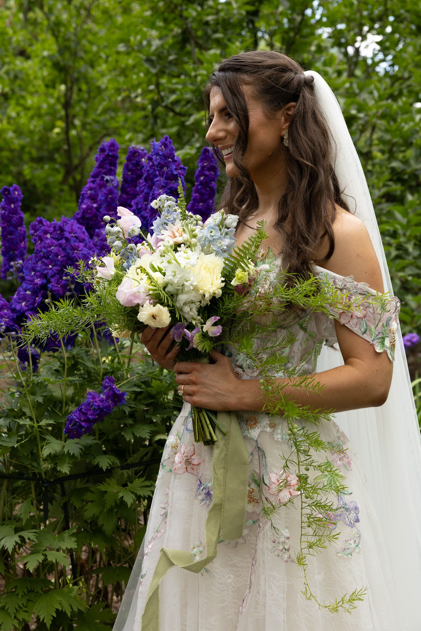 Bride in floral off-shoulder gown holds stunning bouquet with purple delphiniums, white flowers and trailing greenery in garden.