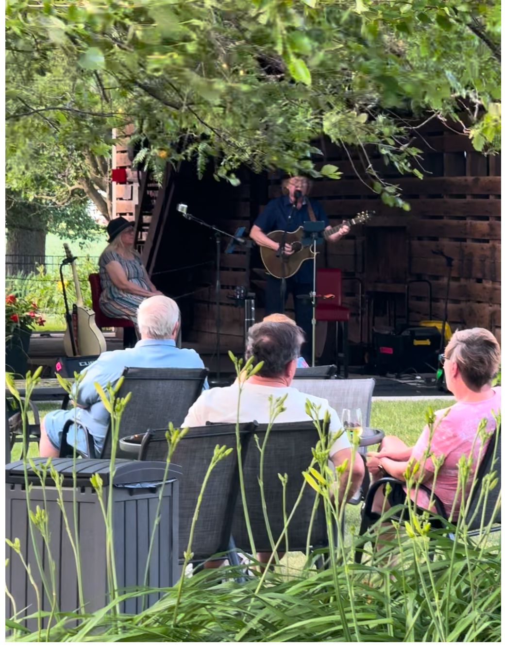 An outdoor concert performance with musicians playing for seated audience members surrounded by lush greenery.