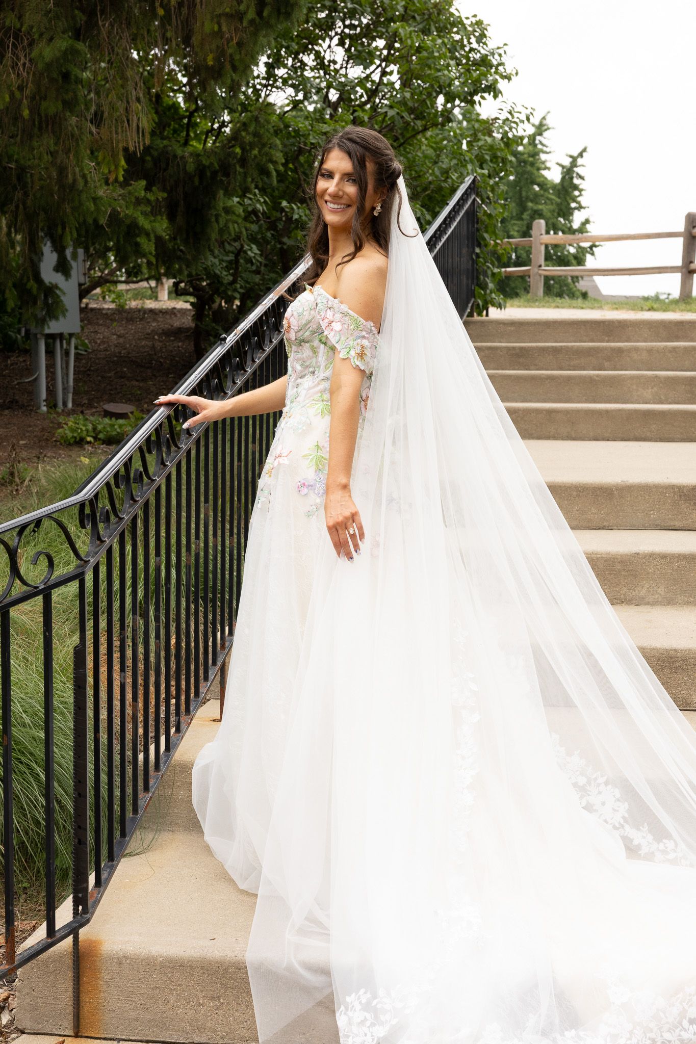 A bride in a flowing white wedding dress and veil gracefully poses on concrete steps with an ornate black railing.
