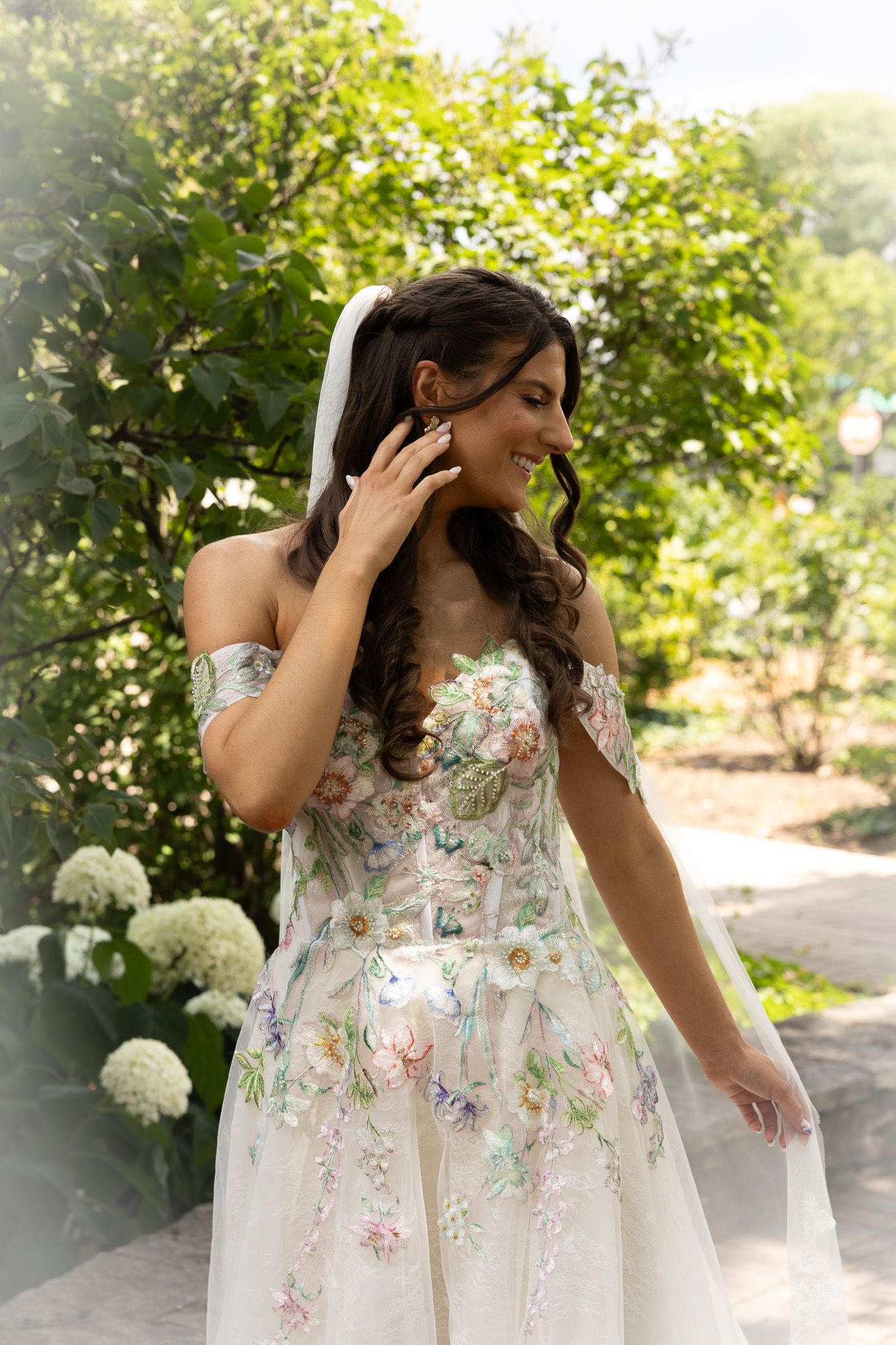 Bride in strapless floral embroidered wedding gown poses outdoors against green foliage and white hydrangeas.
