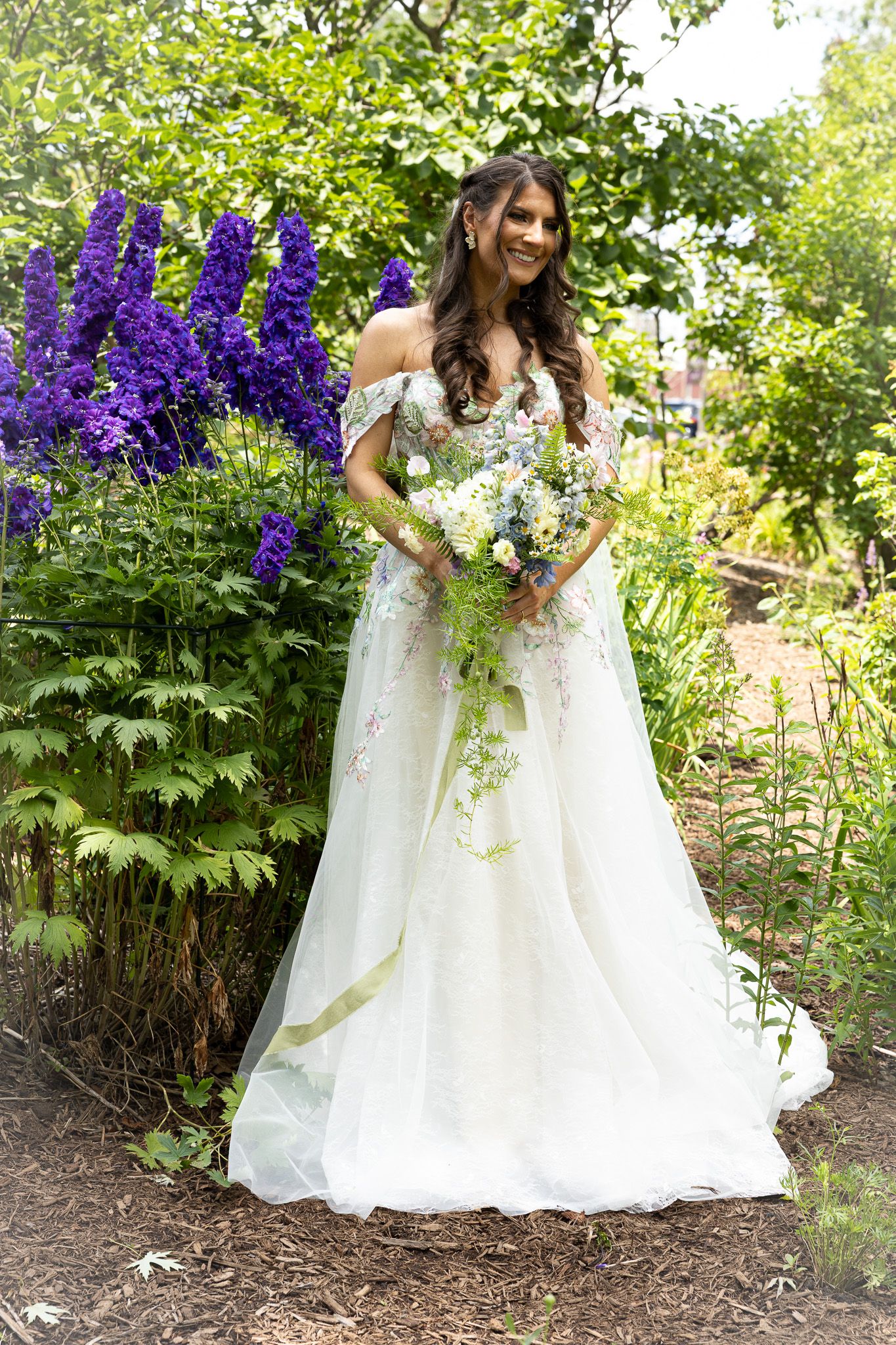 A bridal portrait amongst lush garden greenery and purple delphinium flowers in full bloom.