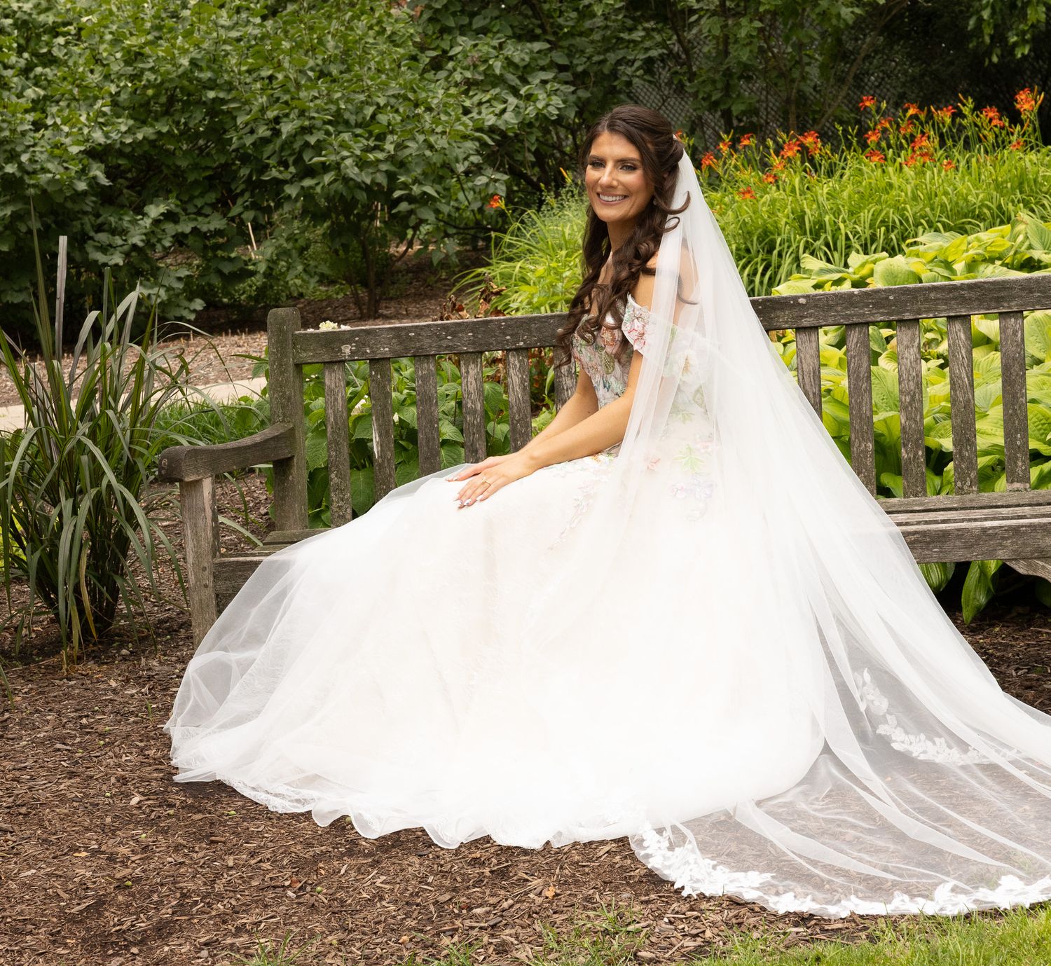 A bride in a flowing white wedding gown sits gracefully on a rustic wooden bench in a lush garden setting.