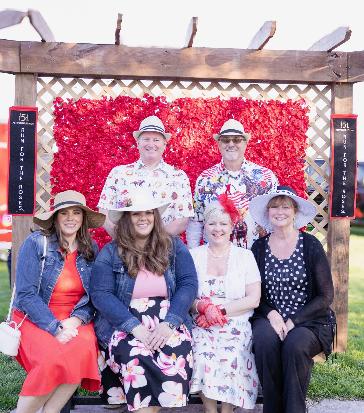 A group poses together in front of a wooden pergola with a vibrant red flower wall backdrop at an outdoor event.