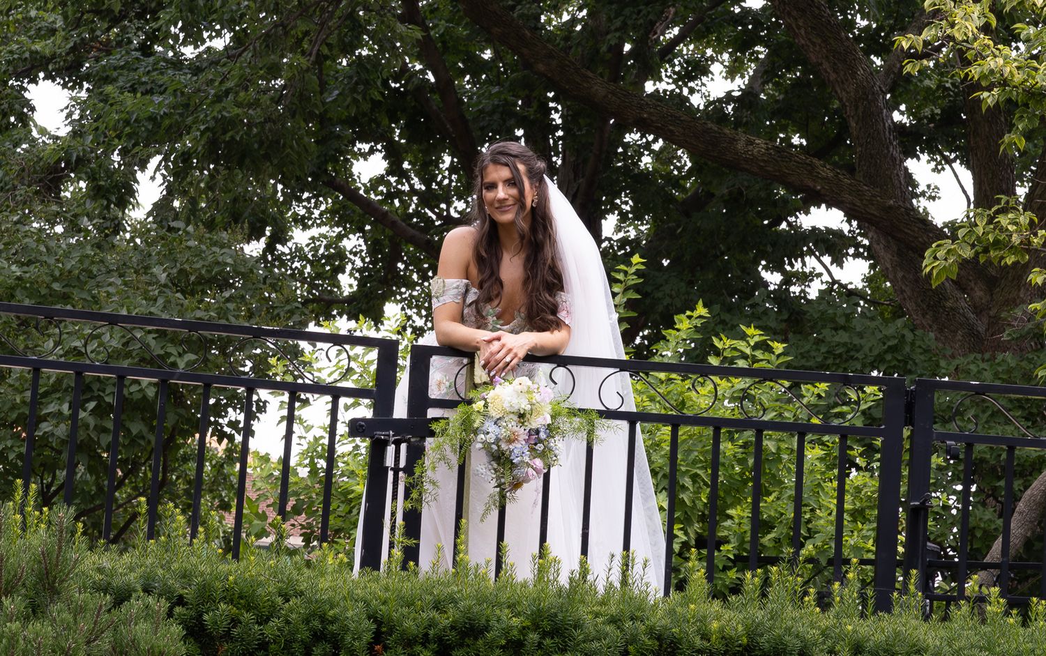 A beautiful bride in a white wedding gown holding a bouquet stands at an ornate iron railing in a tranquil garden setting.
