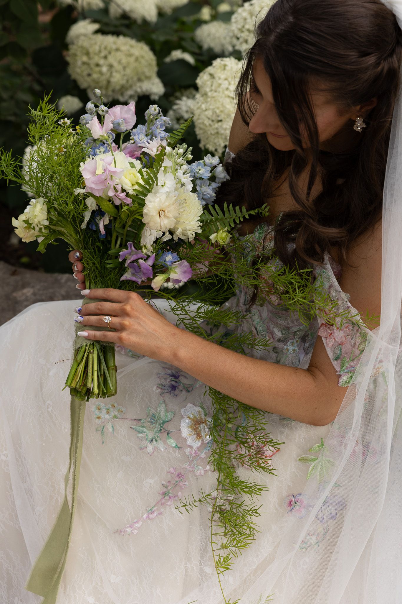 A bride in a white dress admires her beautiful wedding bouquet with white and purple flowers and greenery.