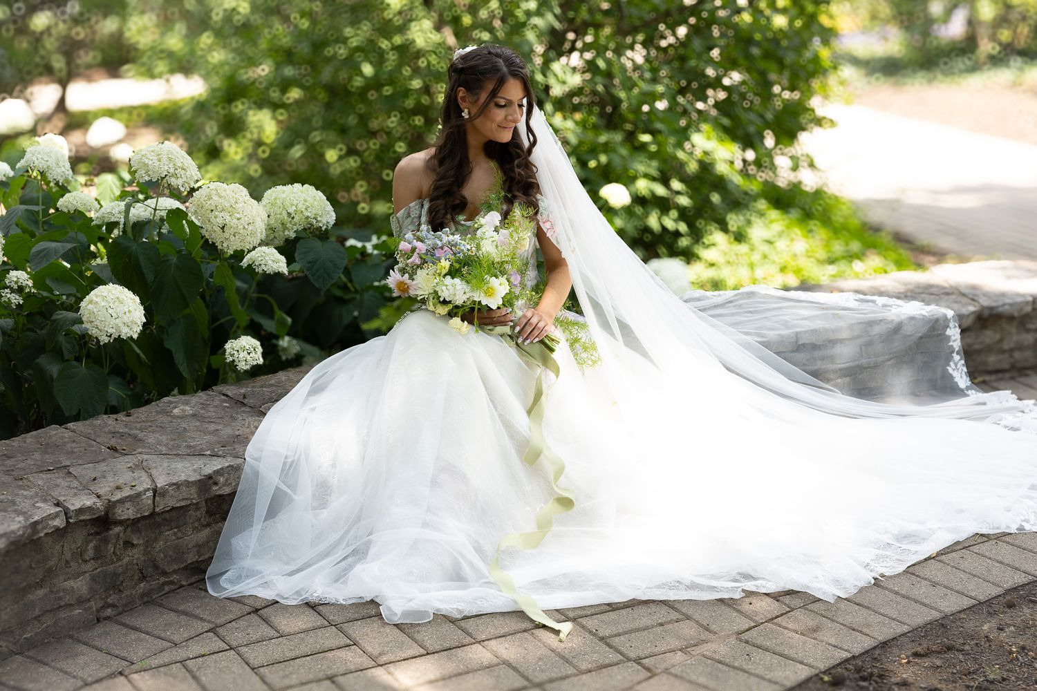 A bride in a flowing white wedding dress poses gracefully on a stone wall surrounded by lush summer greenery.