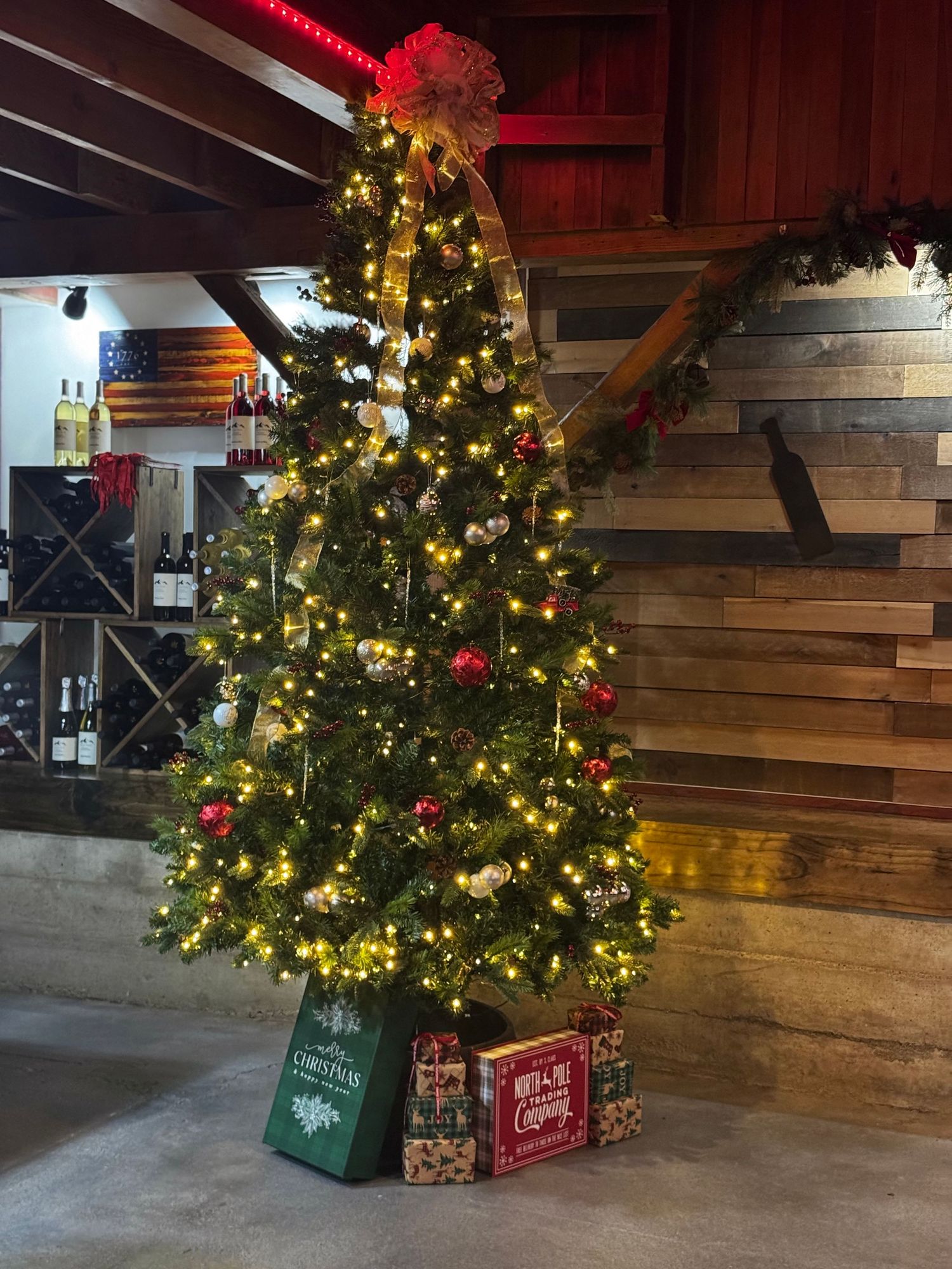 A decorated Christmas tree with twinkling lights and red ornaments stands against a rustic wooden wall with wrapped presents below.