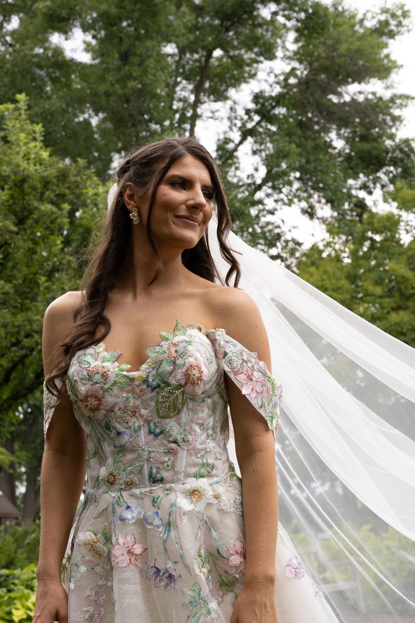An off-shoulder floral wedding dress with delicate embroidery and a flowing veil against a green outdoor backdrop.